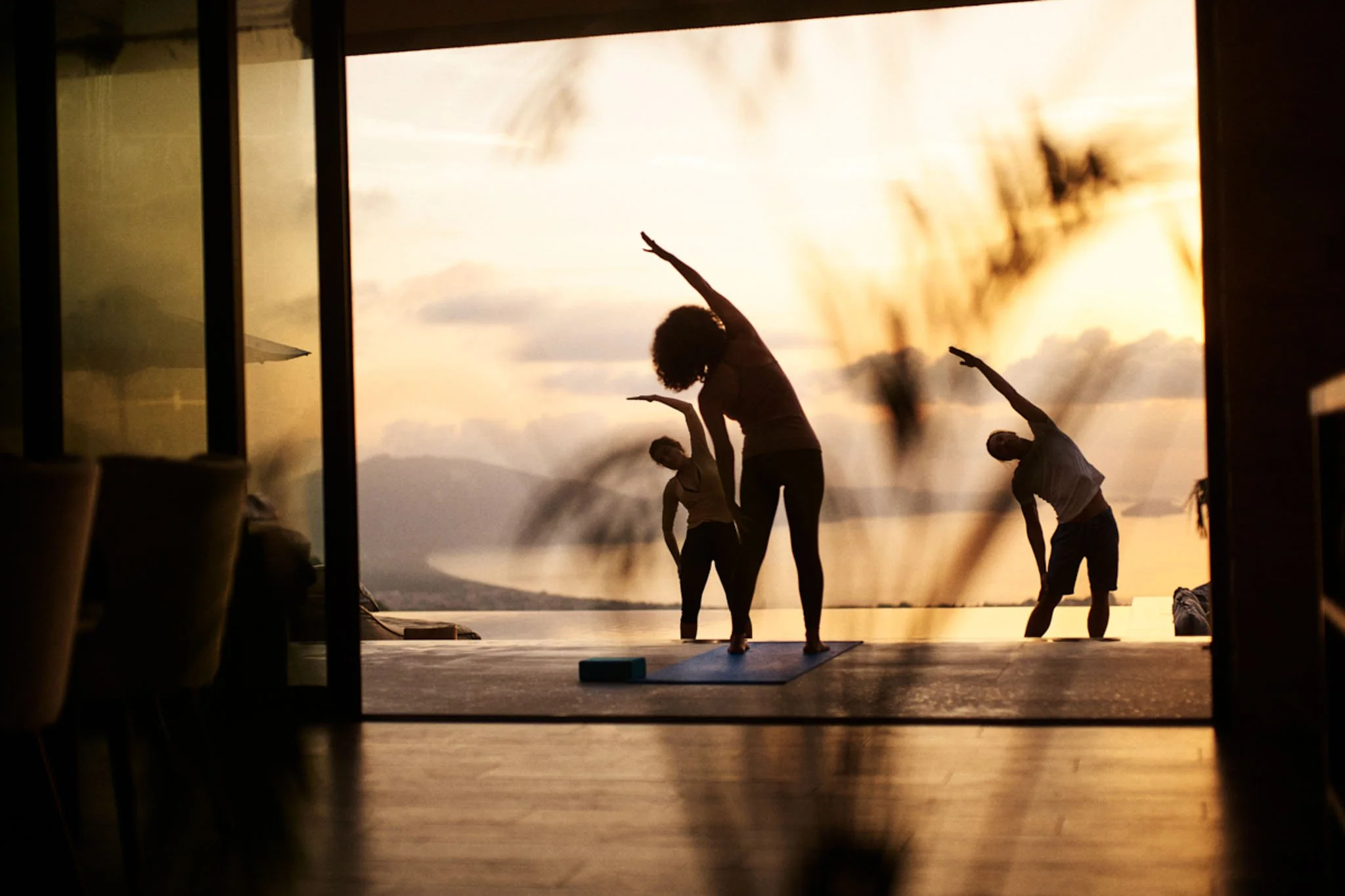 Three people practicing yoga on a patio during sunset, with a scenic view of mountains and water in the background.