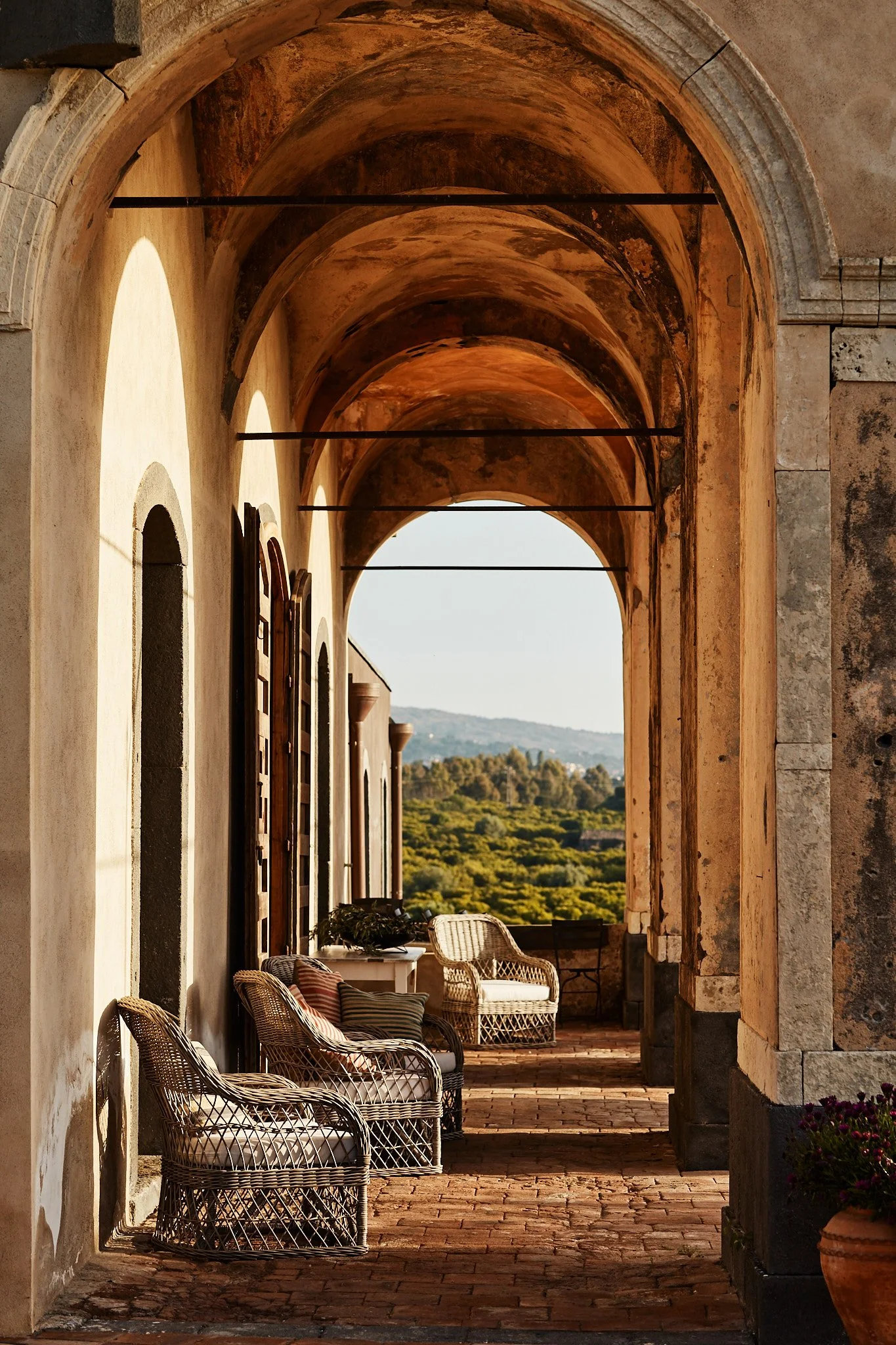 A covered outdoor patio with wicker chairs and cushions, arched ceiling, stone walls, and a scenic view of mountains and lush greenery in the background.