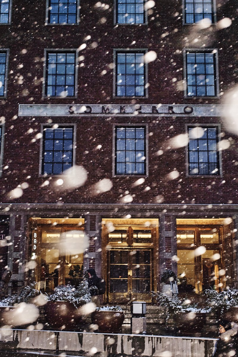 Snow falling in front of a brick building with multiple windows and golden entrance doors, illuminated interior, and a sign that reads 'SOMMERRO' on the facade.