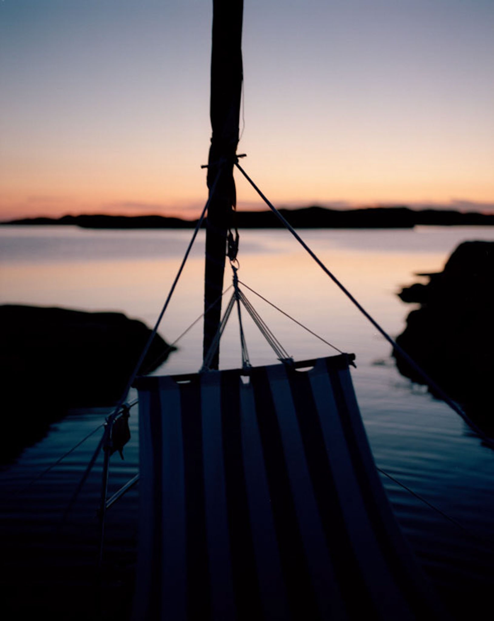 Silhouette of a small sailboat with a striped sail on calm water at sunset, with distant landforms in the background.