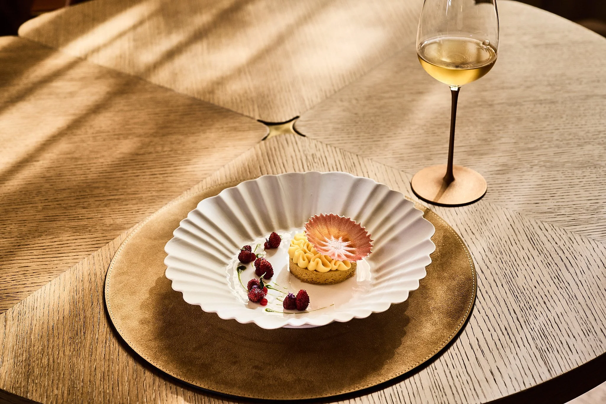 A dessert on a white, scalloped plate with berries and a decorative pink edible flower, accompanied by a glass of white wine on a wooden table with a gold placemat.