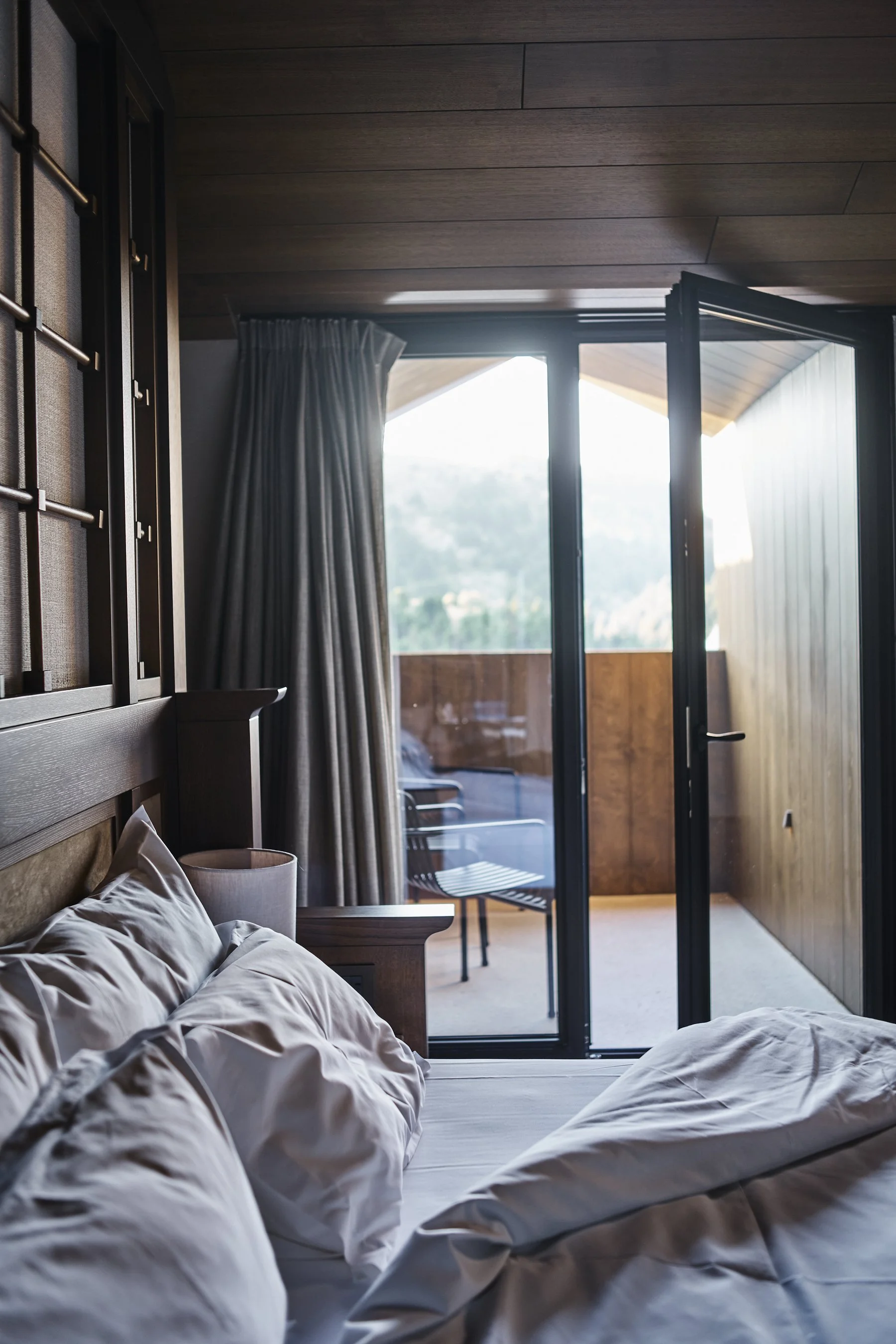A bedroom with unmade bed, pillows, bedside lamp, and access to a balcony with chairs, wooden railings, curtains, and mountain view outside.