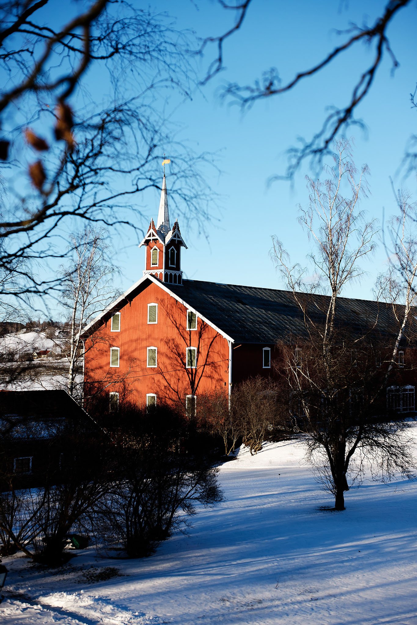 A red wooden church with a tall steeple and a black roof, surrounded by leafless trees on a snowy landscape under a clear blue sky.