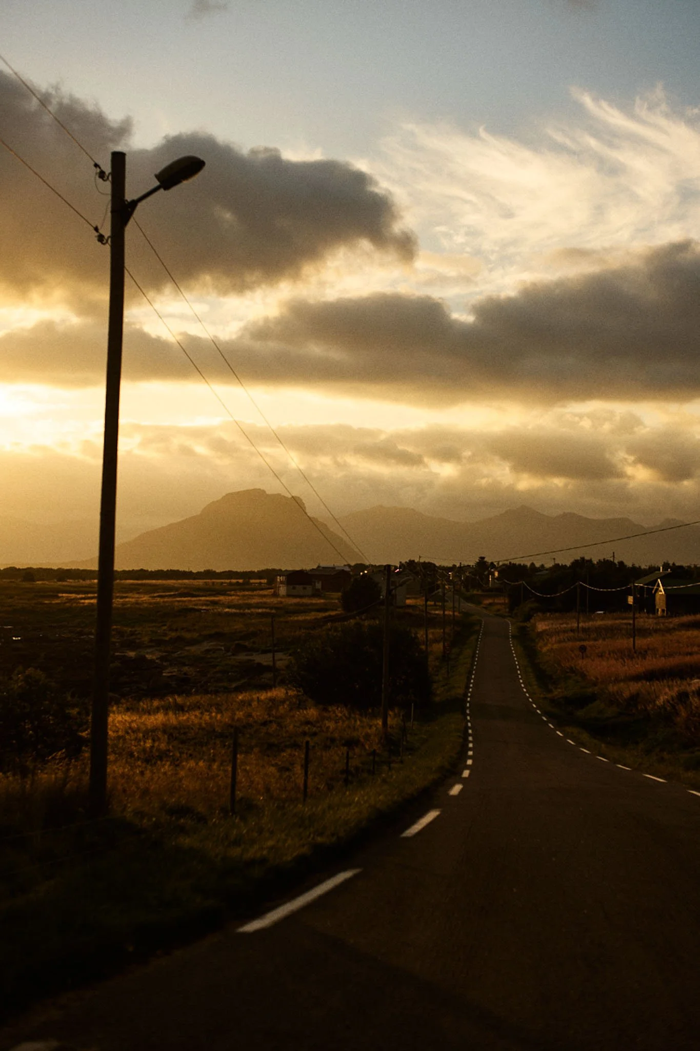 A rural road at sunset with a mountain in the background, utility poles along the roadside, and a partly cloudy sky.