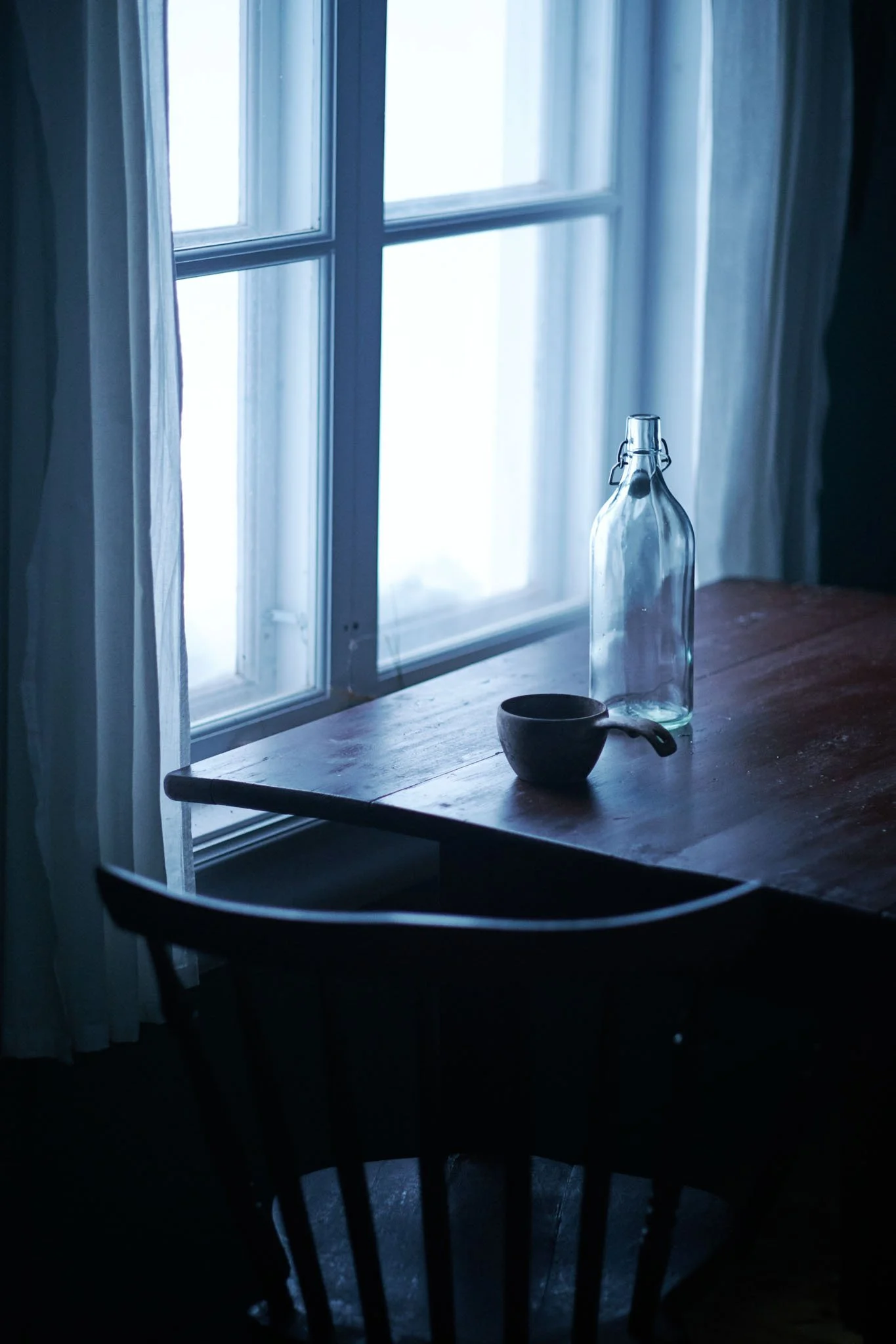 A wooden table near a window with sheer curtains. On the table, there is a glass bottle and a small cup. The scene is dimly lit, with natural light coming through the window.
