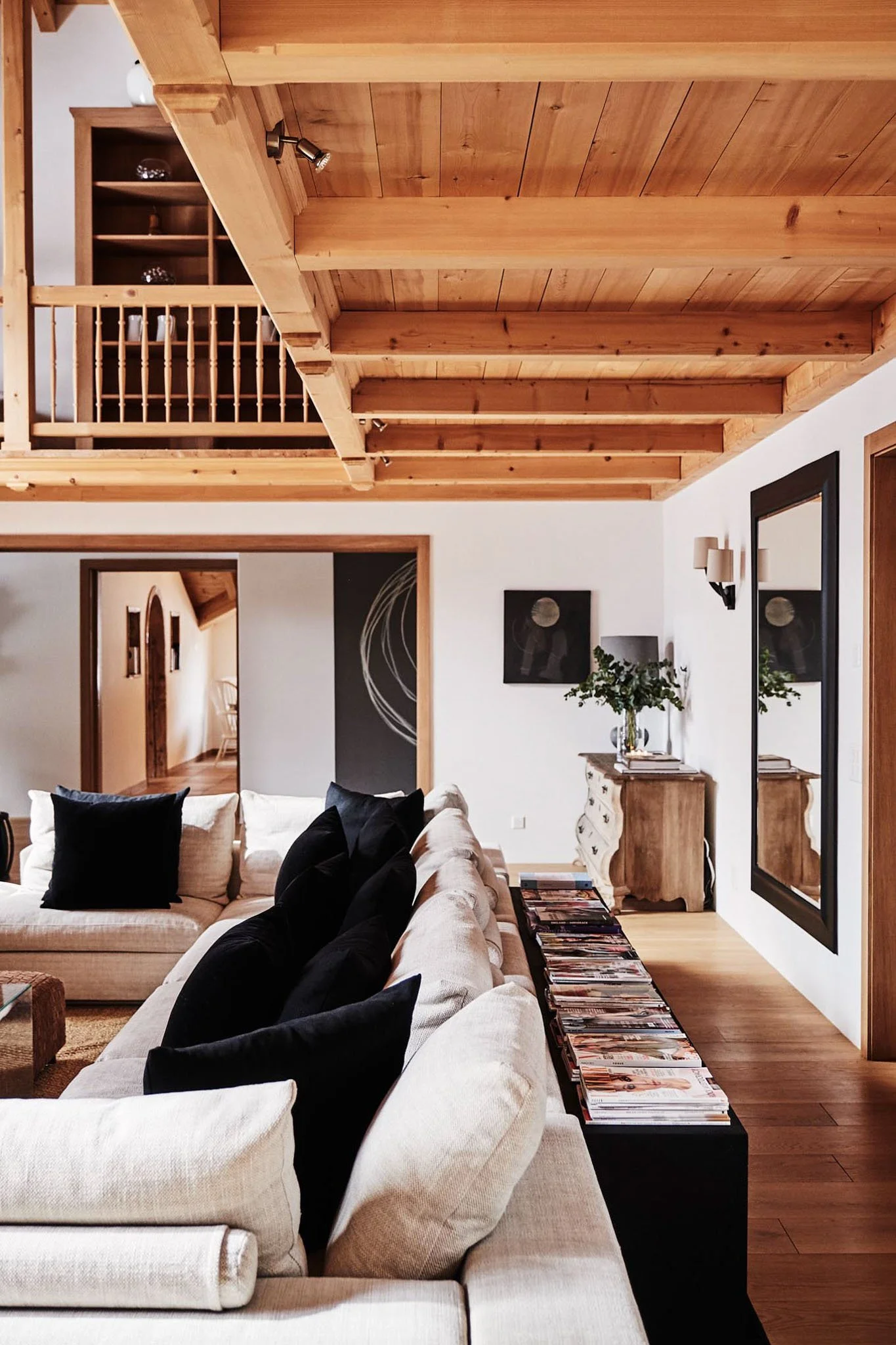 A cozy living room with a beige sectional sofa adorned with black cushions, a wooden coffee table, a black console table filled with magazines, a vintage wooden sideboard with a vase of greenery, a large mirror, wall art, and a wooden ceiling with tr
