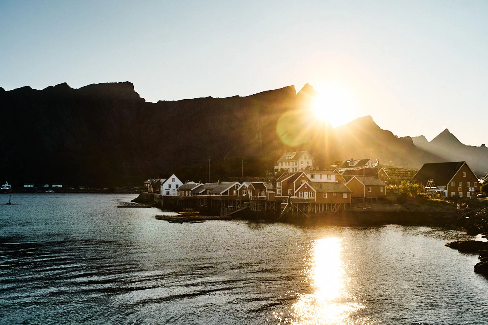 Sunset over a small coastal village with houses on stilts by the water, mountains in the background, and the sun low on the horizon creating a reflection on the water.