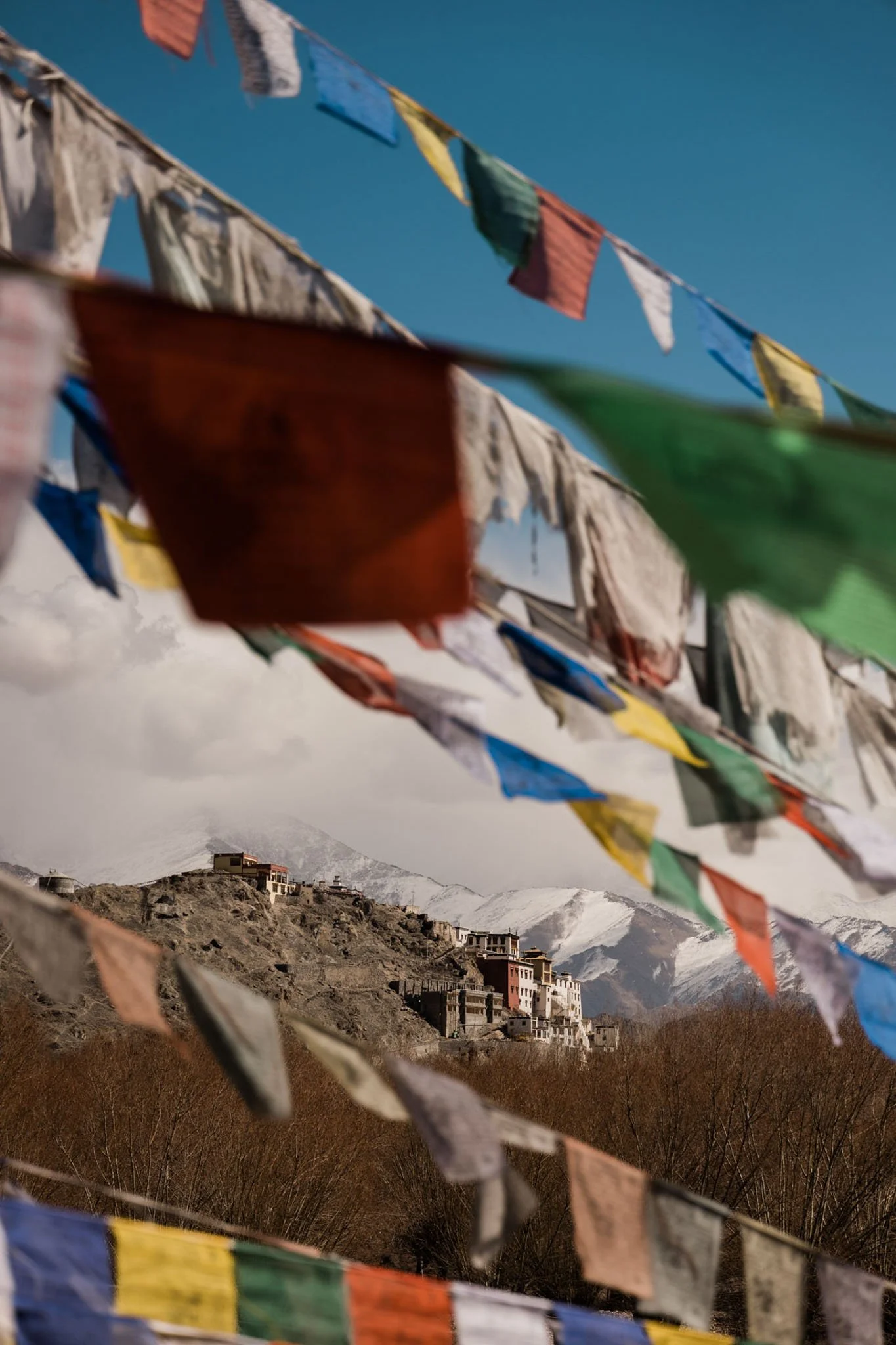 Colorful prayer flags fluttering in the wind against a mountainous landscape with snow-covered peaks and a village of buildings on a hillside.