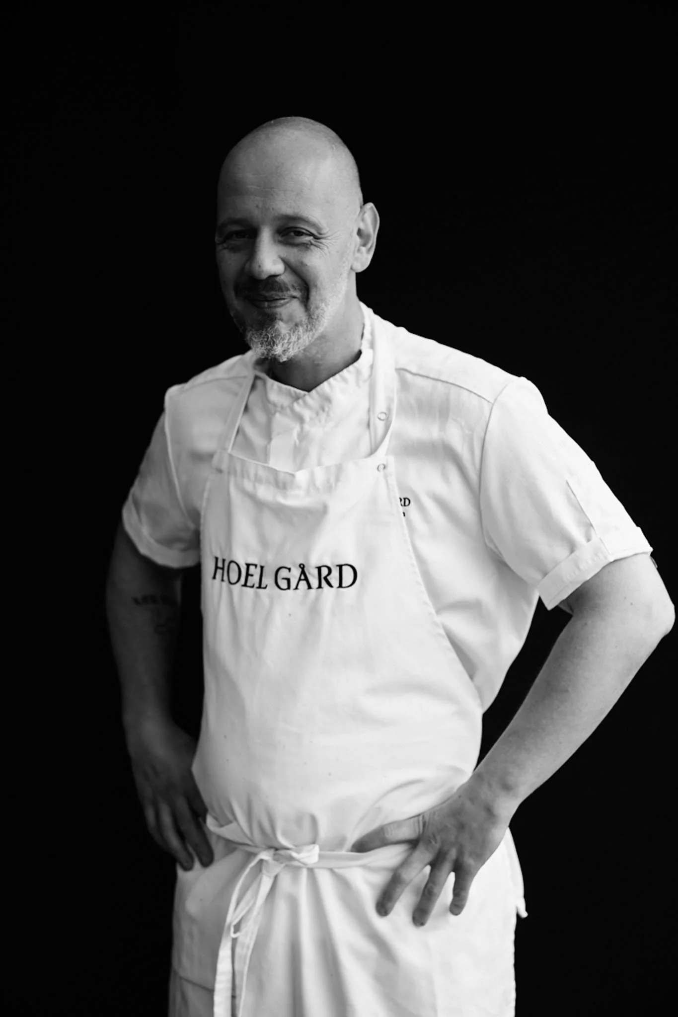 Black and white photo of a smiling male chef with a shaved head and beard, wearing a white chef's coat and apron with "HOELGÅRD" written on it, standing against a dark background.