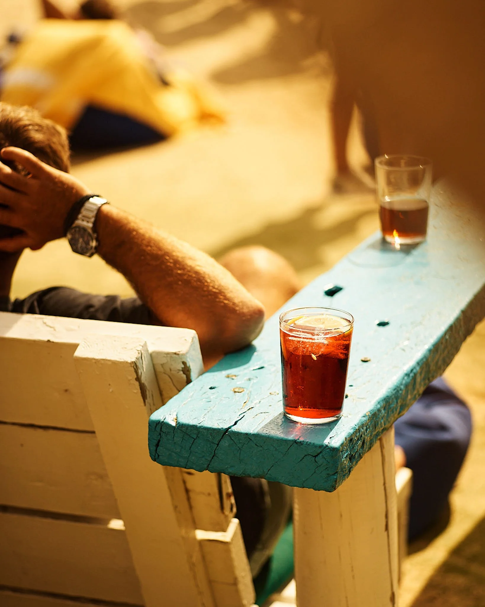 Two glasses of dark-colored drinks on a weathered, painted wooden bench outdoors during nighttime, with a person sitting nearby and a temporary tent in the background.