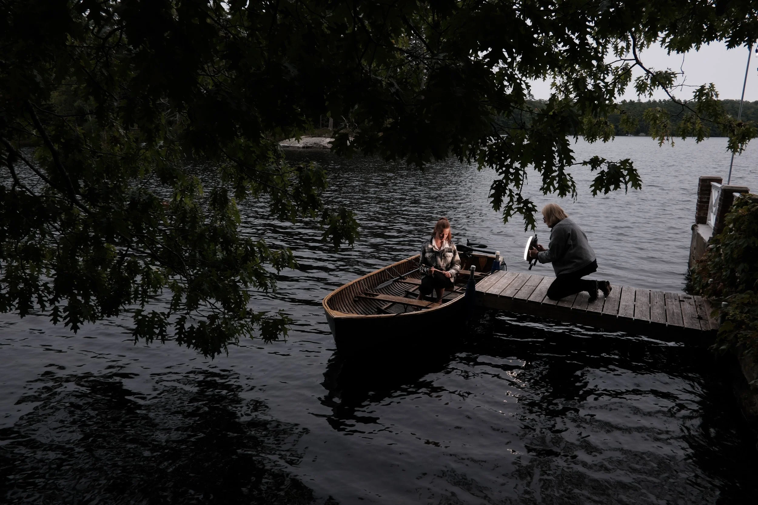 A student photographing a model in a vintage boat on a lake, during a Greg Gorman workshop at Blenheim Park Maine, on a dock next to a small boat on a lake, with one woman sitting inside the boat and the other woman kneeling on the dock.