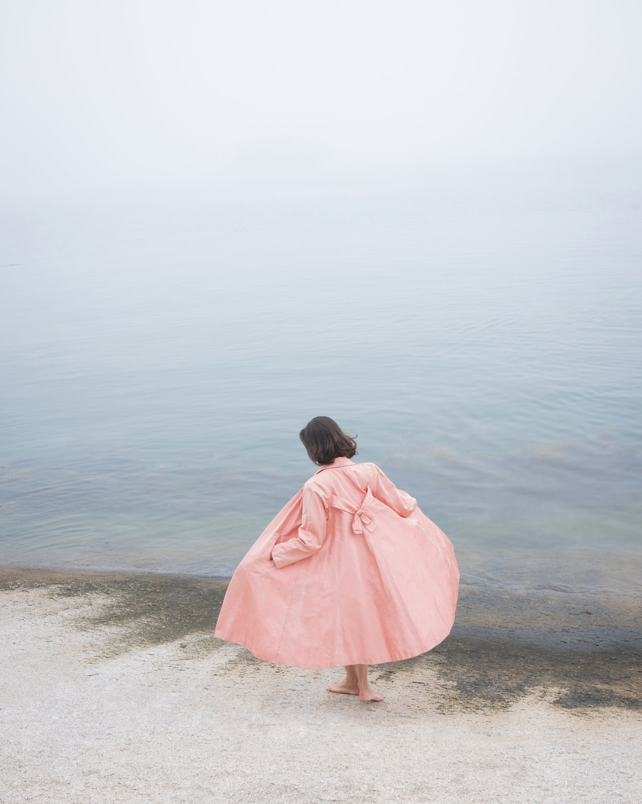 A Cig Harvey photograph featuring a person wearing a pink dress standing barefoot on a sandy beach, looking at the water.