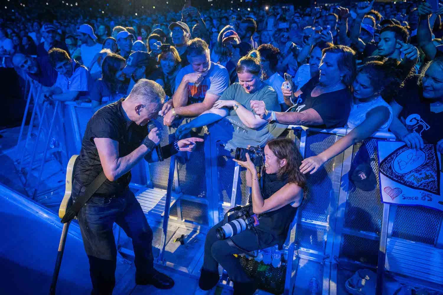 A Rob DeMartin photograph of Bruce Springsteen interacting with his sister, photographer Pamela Springsteen at a concert.  Behind them fans hold signs and cheer.  The scene is illuminated with vibrant blue lighting.