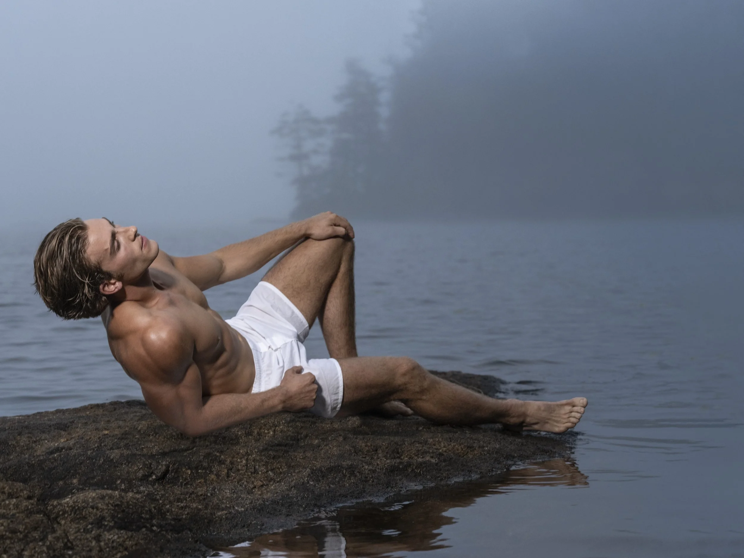 A fine art image made during a Greg Gorman workshop featuring a shirtless man with long hair in white shorts relaxing on a rock by a misty lake, with trees in the background.