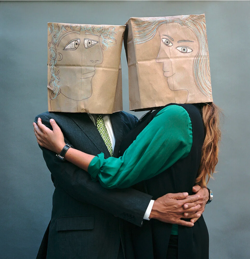 An Aline Smithson photography showing two people wearing paper bags over their heads with hand-drawn faces, embracing each other.