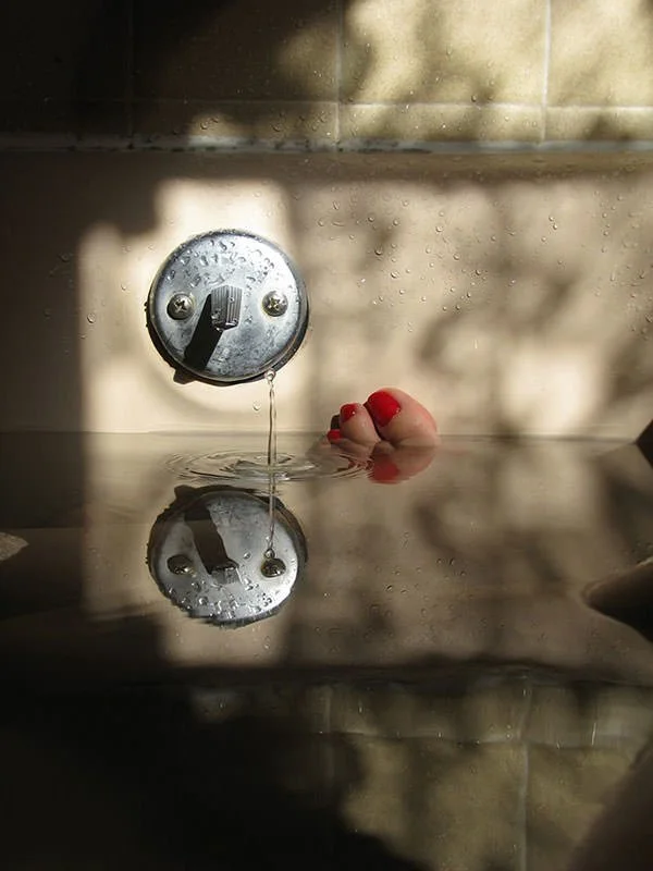 A Maggie Steber image showing a close-up of a shower drain with a single water droplet falling and creating ripples, reflecting the drain on the surface below, with a person's foot with bright red nail polish visible in the background.