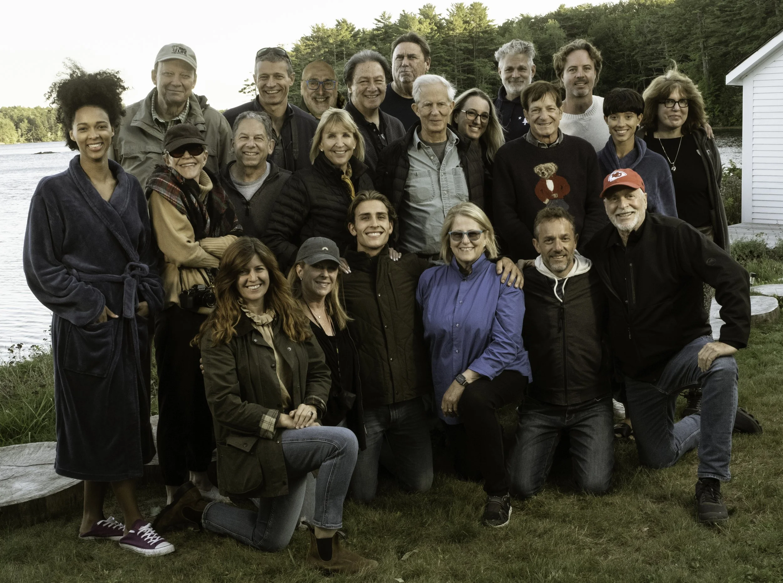 A group of about 20 Blenheim Park Maine students gathered outdoors near a body of water, smiling for a photo during daytime with trees and a white house in the background.