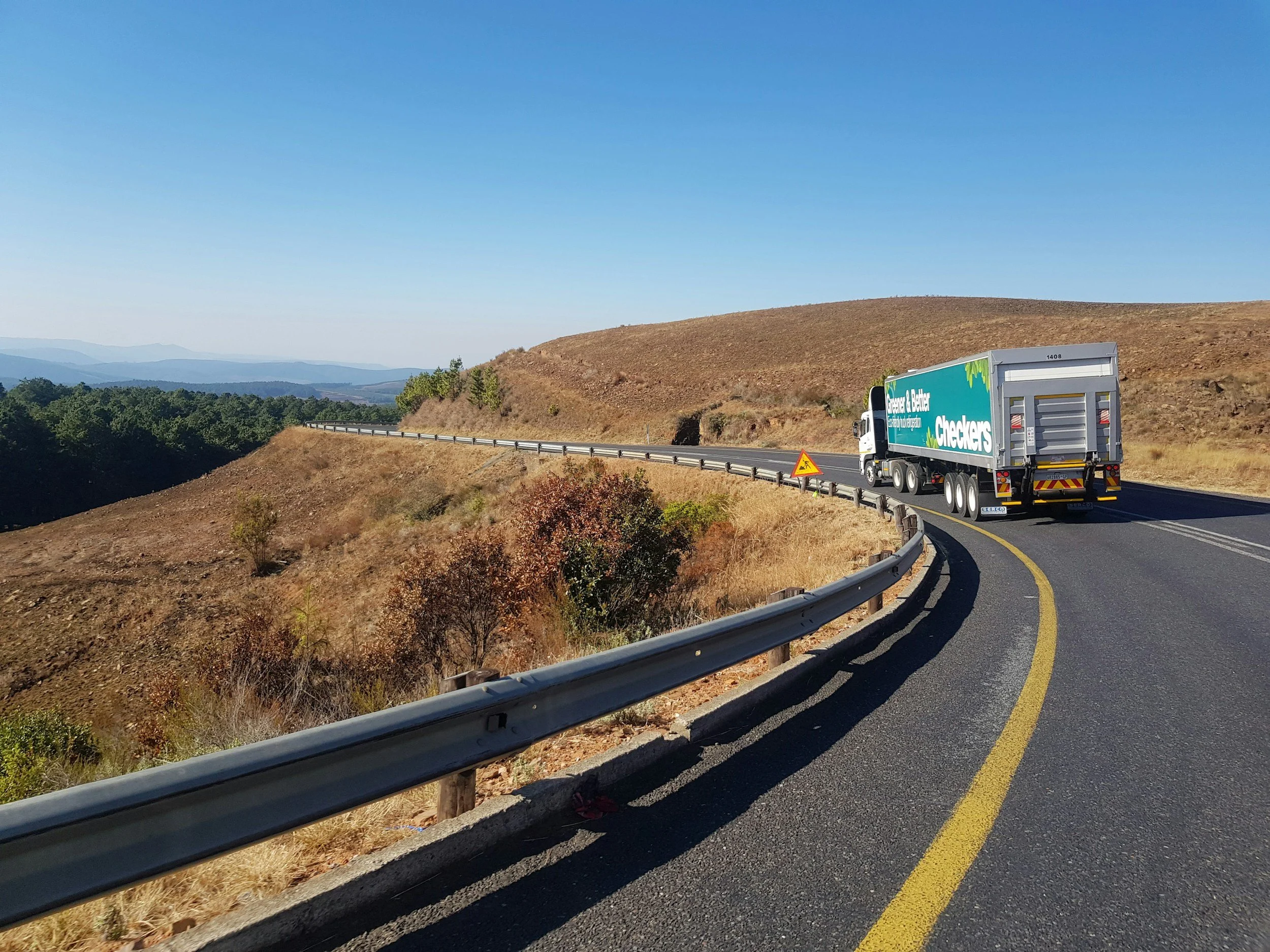 Un camion roulant sur une route sinueuse dans un paysage de collines sèches et herbeuses, avec un ciel clair et dégagé.