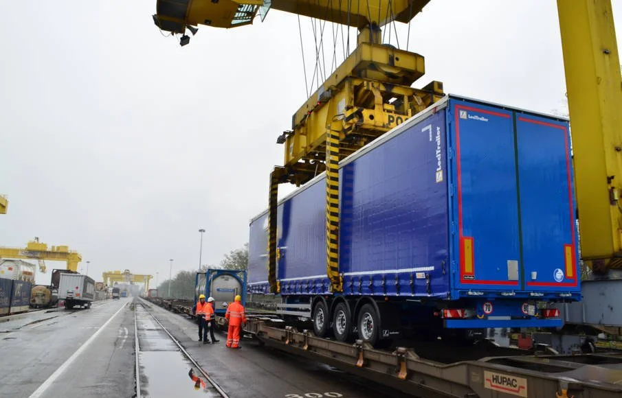 Gros plan sur un camion bleu chargé sur un wagon dans un terminal ferroviaire, avec des travailleurs en vêtements de sécurité orange à proximité, sous un ciel nuageux.