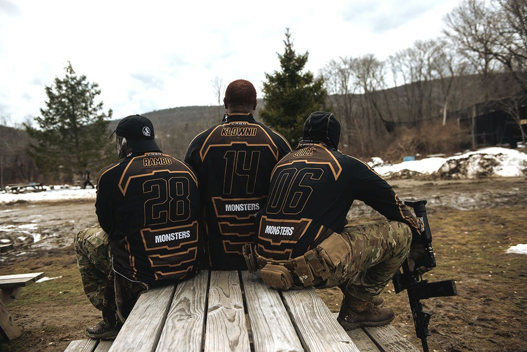 Three individuals wearing black and gold jerseys with the word 'Monsters' and numbers, sitting on a wooden bench outdoors with trees, snow, and cloudy sky in the background.
