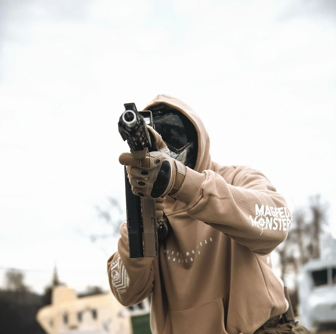 A person wearing a hoodie and gloves, aiming a handgun outdoors against a cloudy sky.