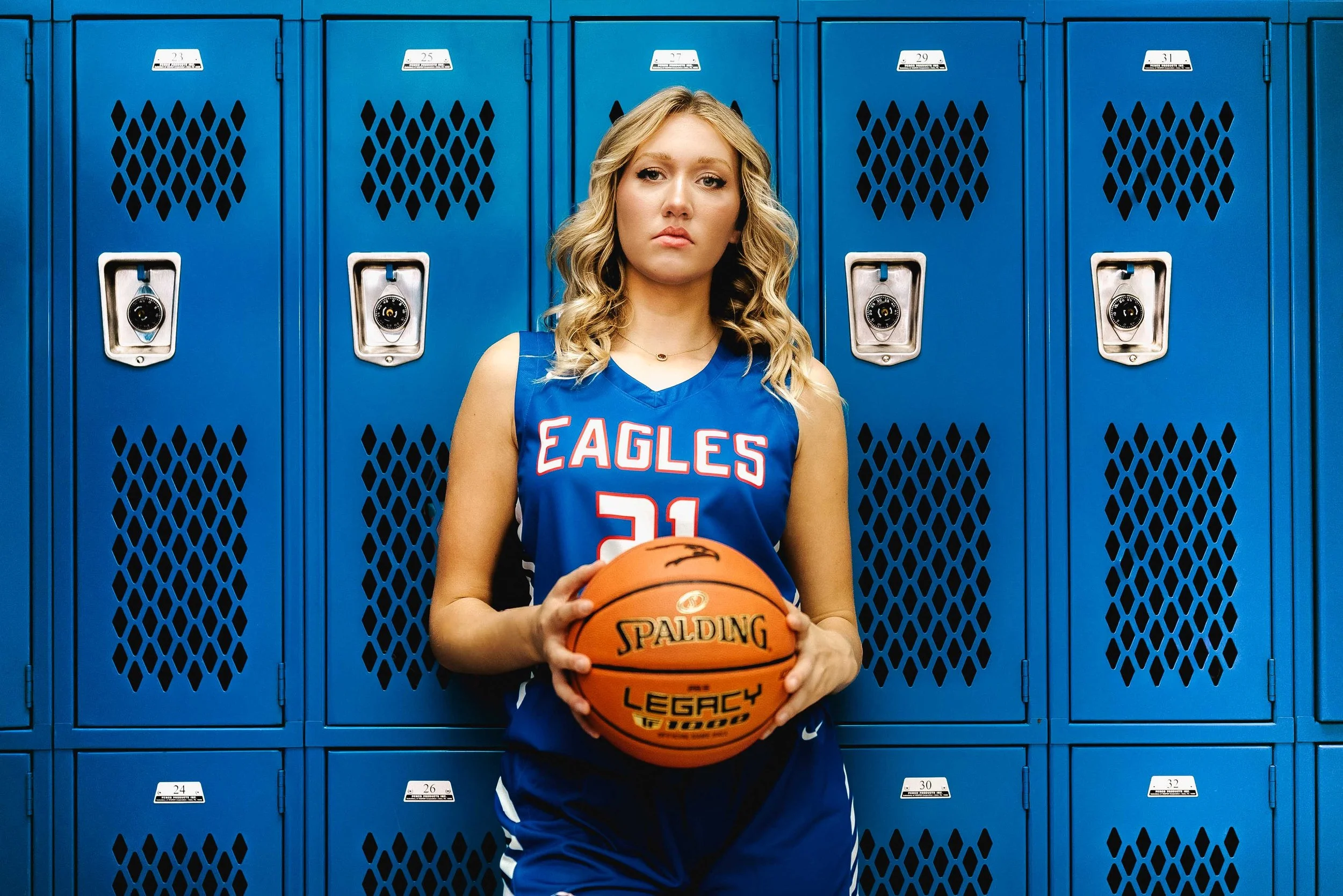 A young female basketball player standing in front of blue gym lockers, holding a basketball. She is wearing a blue jersey with 'EAGLES' and the number 21 on it.