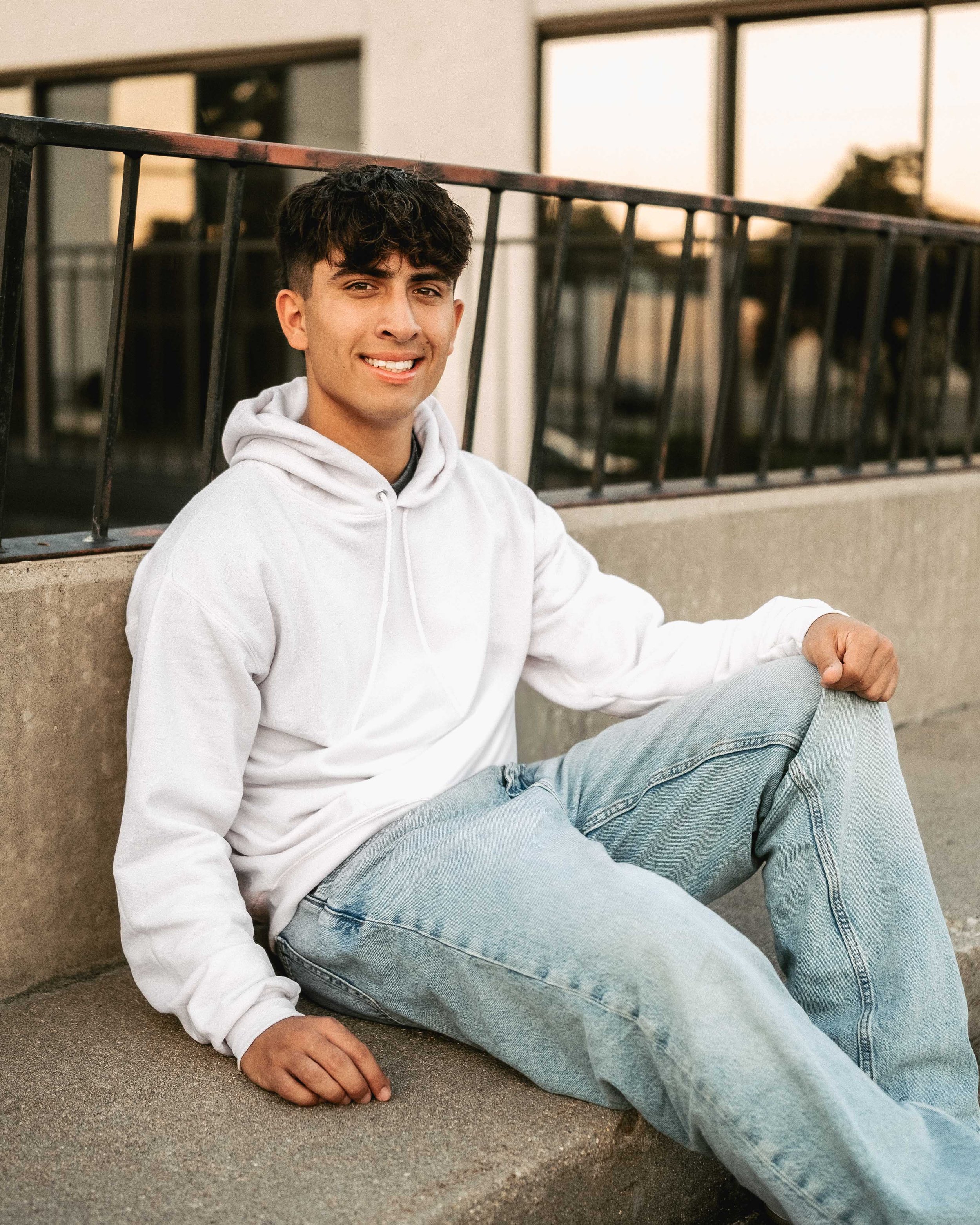 A young man with dark, curly hair and light skin, sitting on the ground outside near a building, wearing a white hoodie and light-wash jeans, smiling at the camera during sunset.
