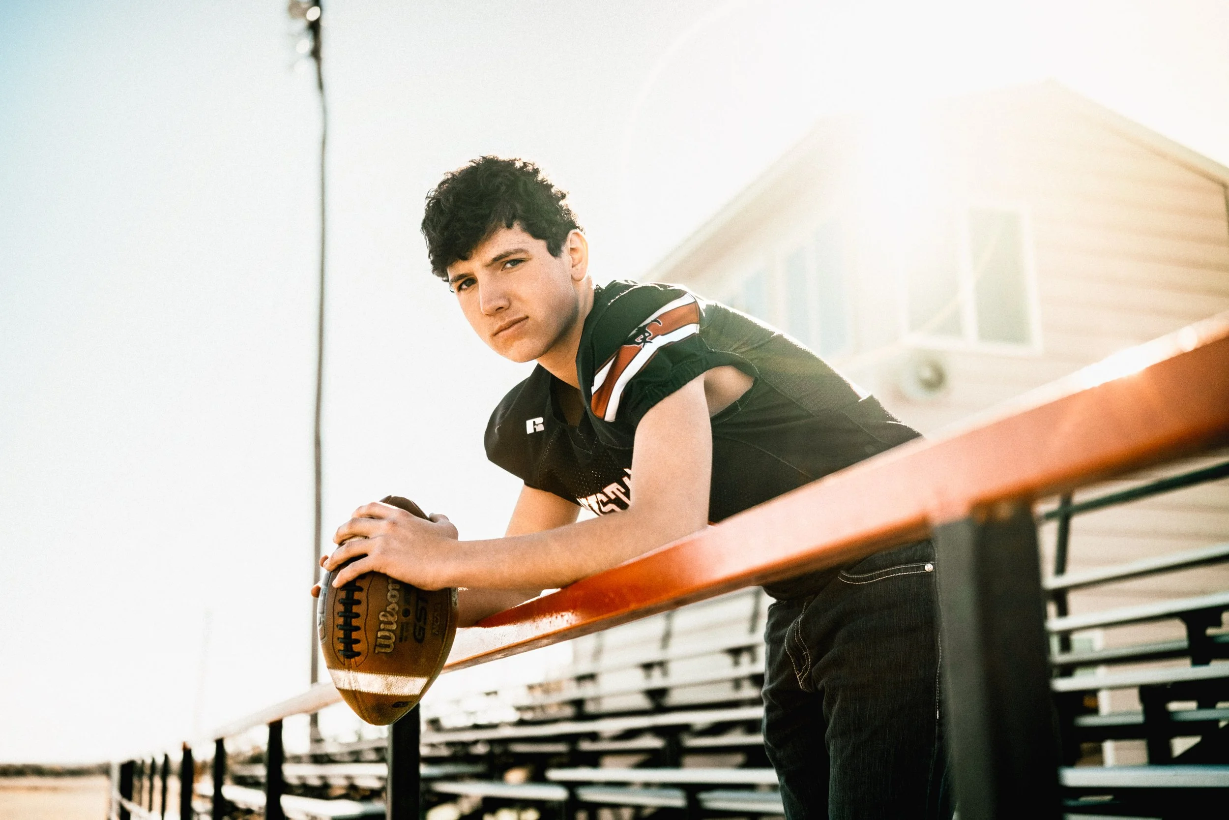 A teenage boy wearing a black football jersey leans on a red and black railing, holding an American football with both hands. He has short dark curly hair and looks into the camera with a serious expression. The background shows a house and a bright 