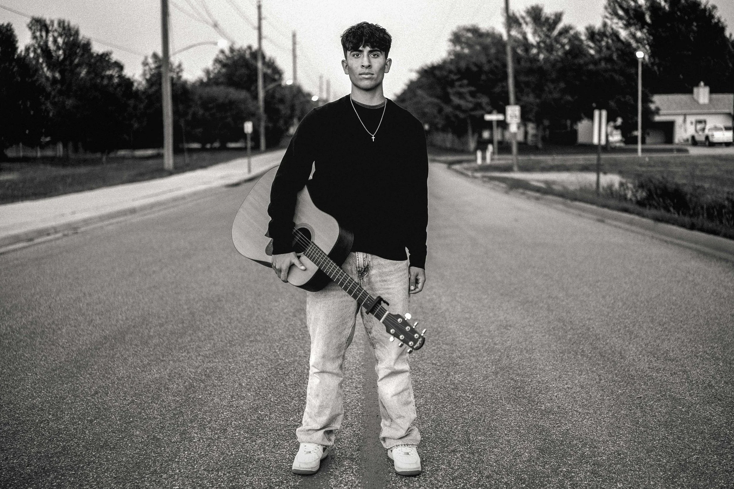 A young man standing in the middle of an empty street holding an acoustic guitar, dressed casually in a black long-sleeve shirt, jeans, and sneakers, with trees and houses in the background in black-and-white.