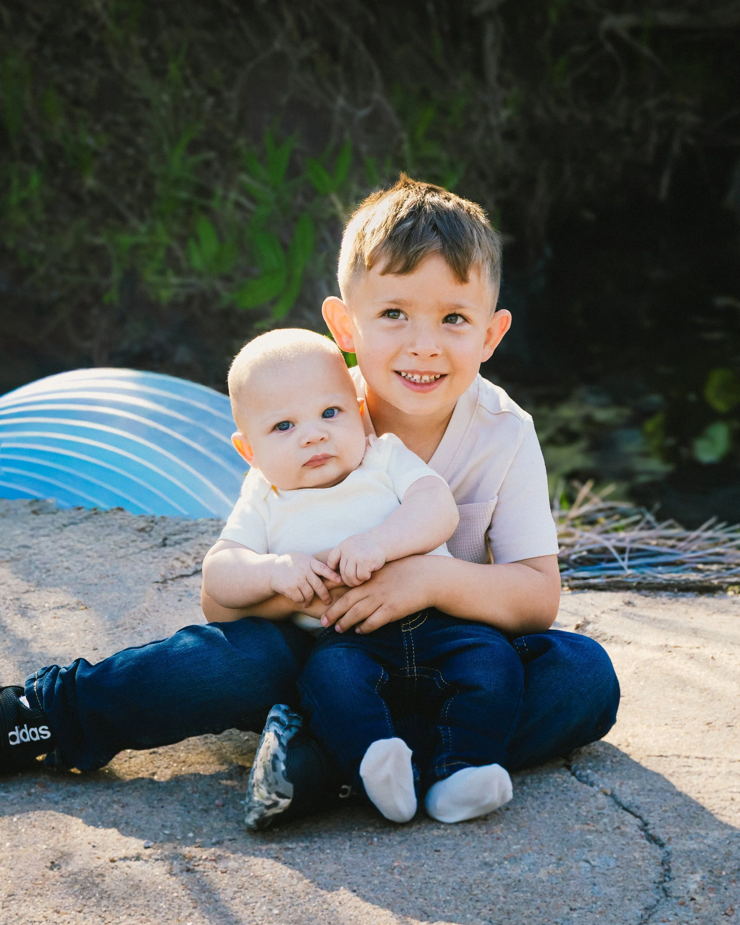 A young boy and a baby sitting on a concrete surface outdoors, with greenery behind them. The boy is smiling and holding the baby, who has a neutral expression.