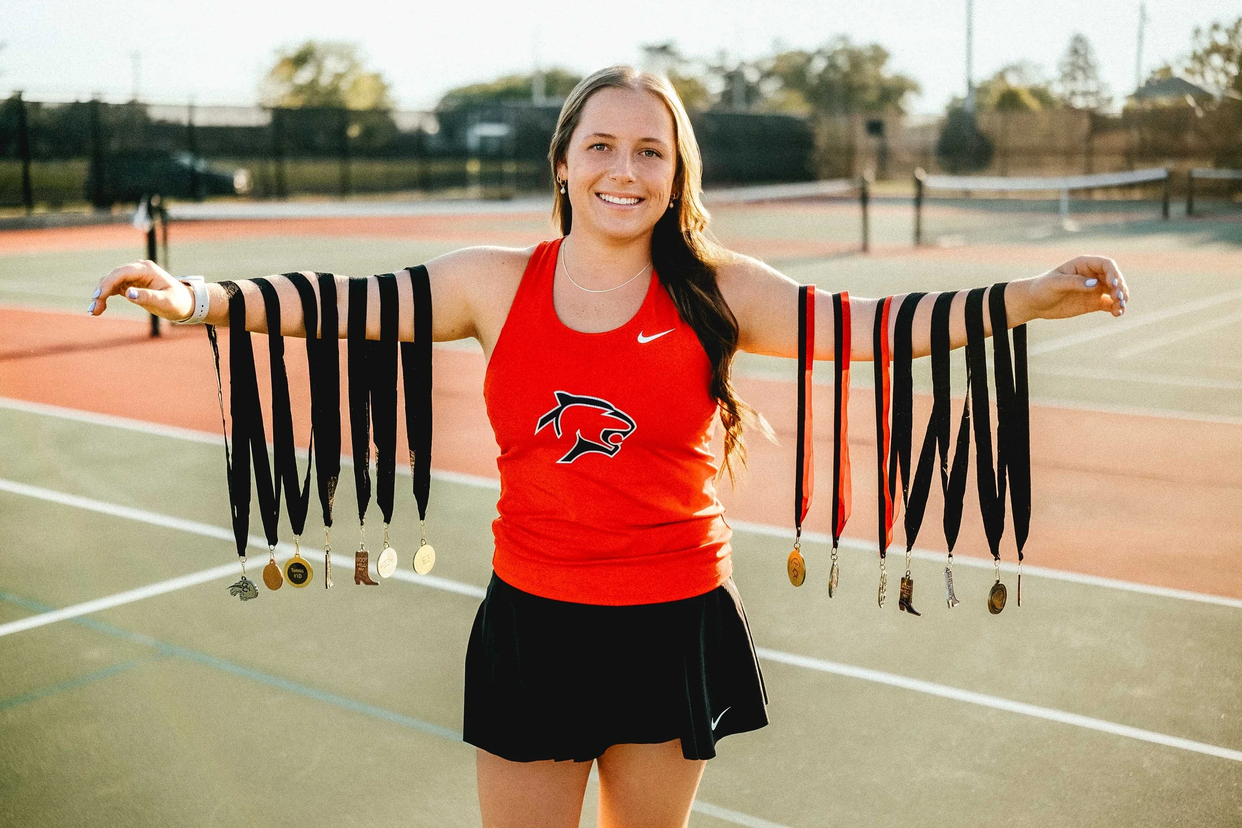 Female athlete standing on a tennis court, holding multiple medals with both arms extended, wearing a red sports top with a panther logo, black shorts, and a smile.