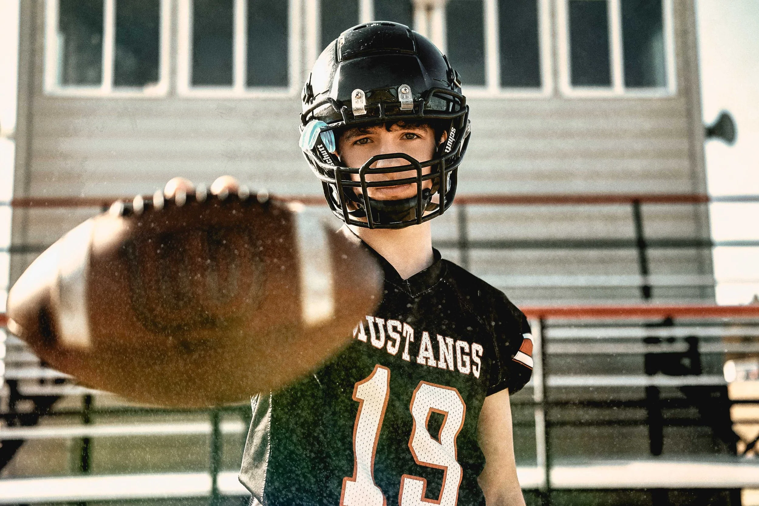 A young football player wearing a black helmet and a black jersey with the number 13 and 'MUSTANGS' written on it, holding a football towards the camera outdoors.