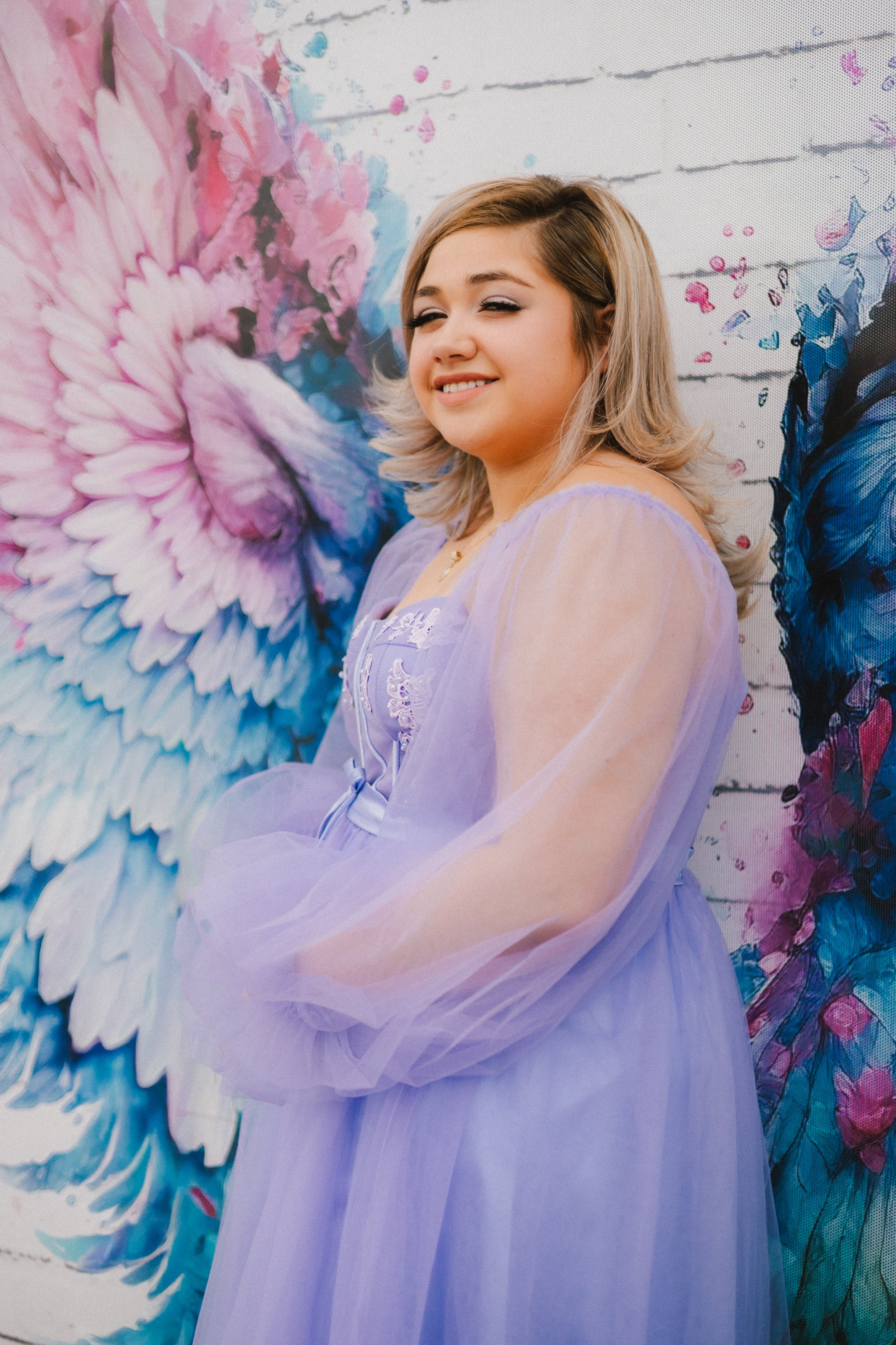 A young woman with blonde hair and light makeup, wearing a lavender dress and sheer matching cape, leaning against a mural of colorful angel wings painted on a white brick wall.