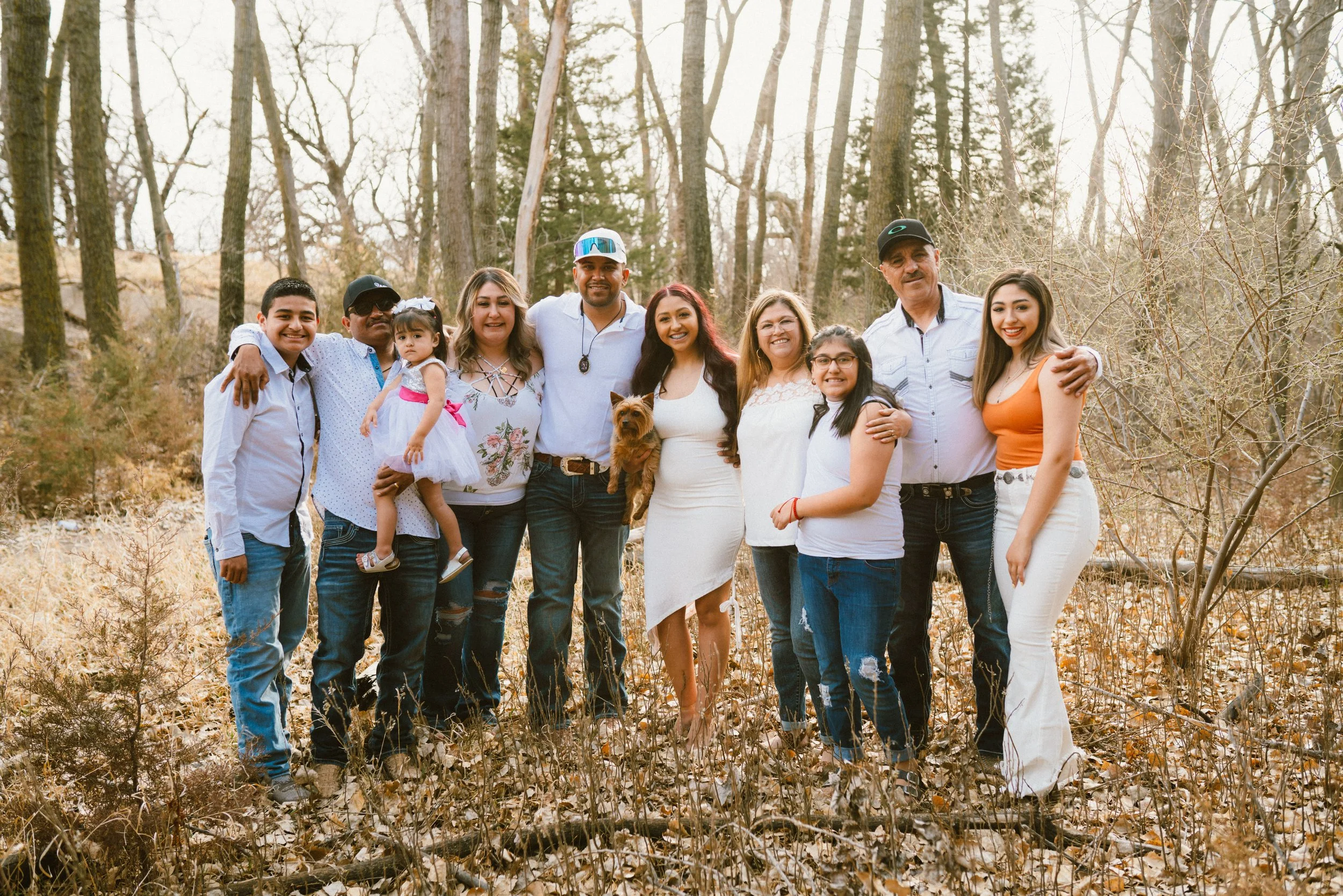 A large, multigenerational family of eleven people standing together in a wooded area with leafless trees and fallen leaves, smiling at the camera, including children, adults, and elders, with one small dog.