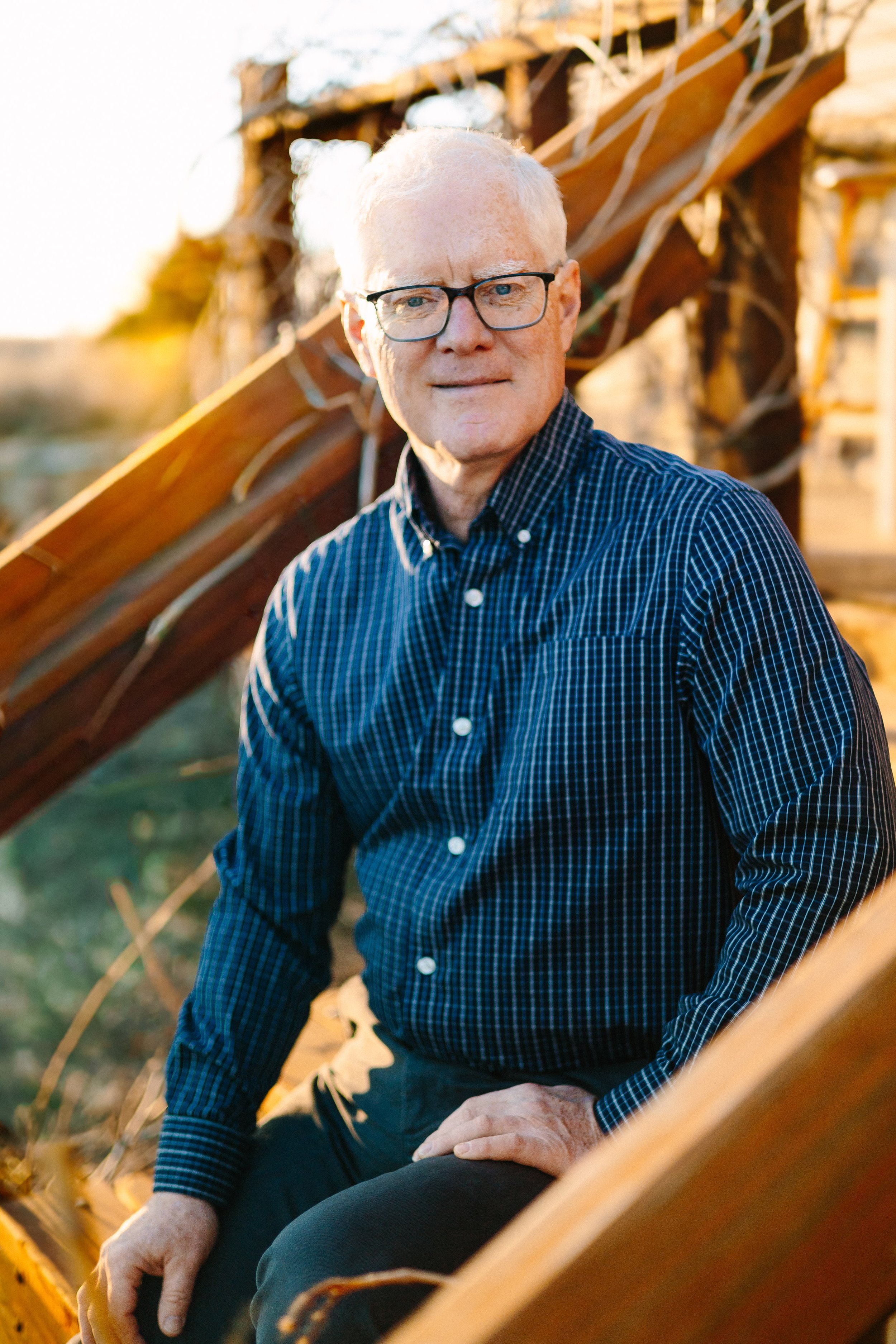 An elderly man with glasses and white hair, wearing a dark checkered shirt and dark pants, sitting on a wooden structure outdoors during sunset.