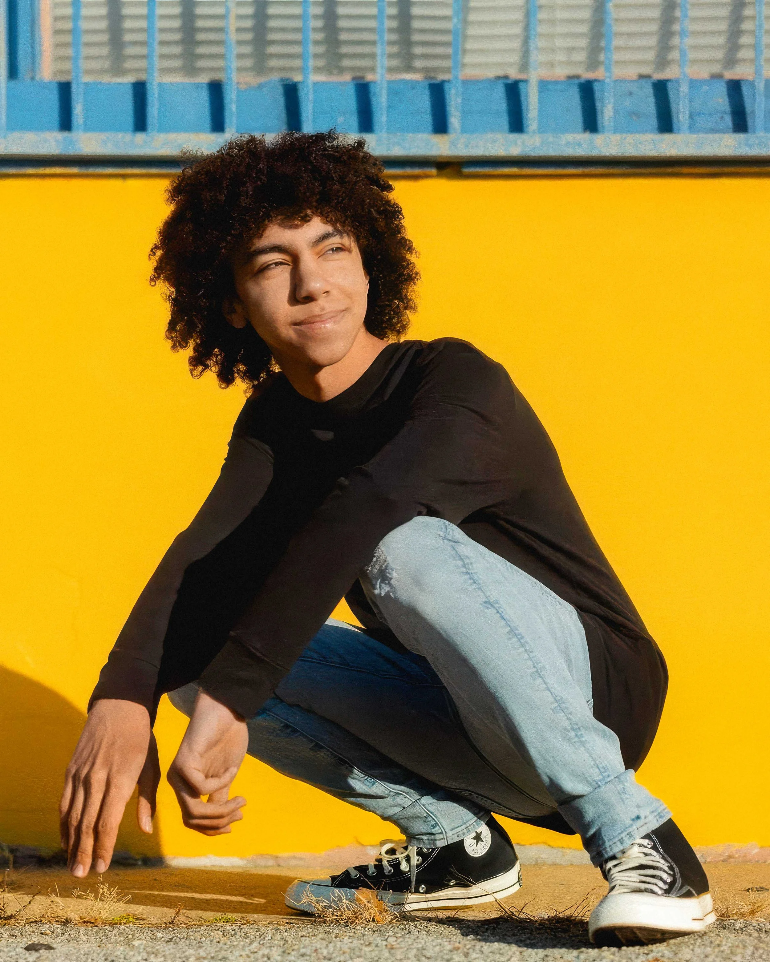 A young man with curly hair wearing a black long-sleeve shirt, light blue jeans, and black Converse sneakers squatting in front of a bright yellow wall.