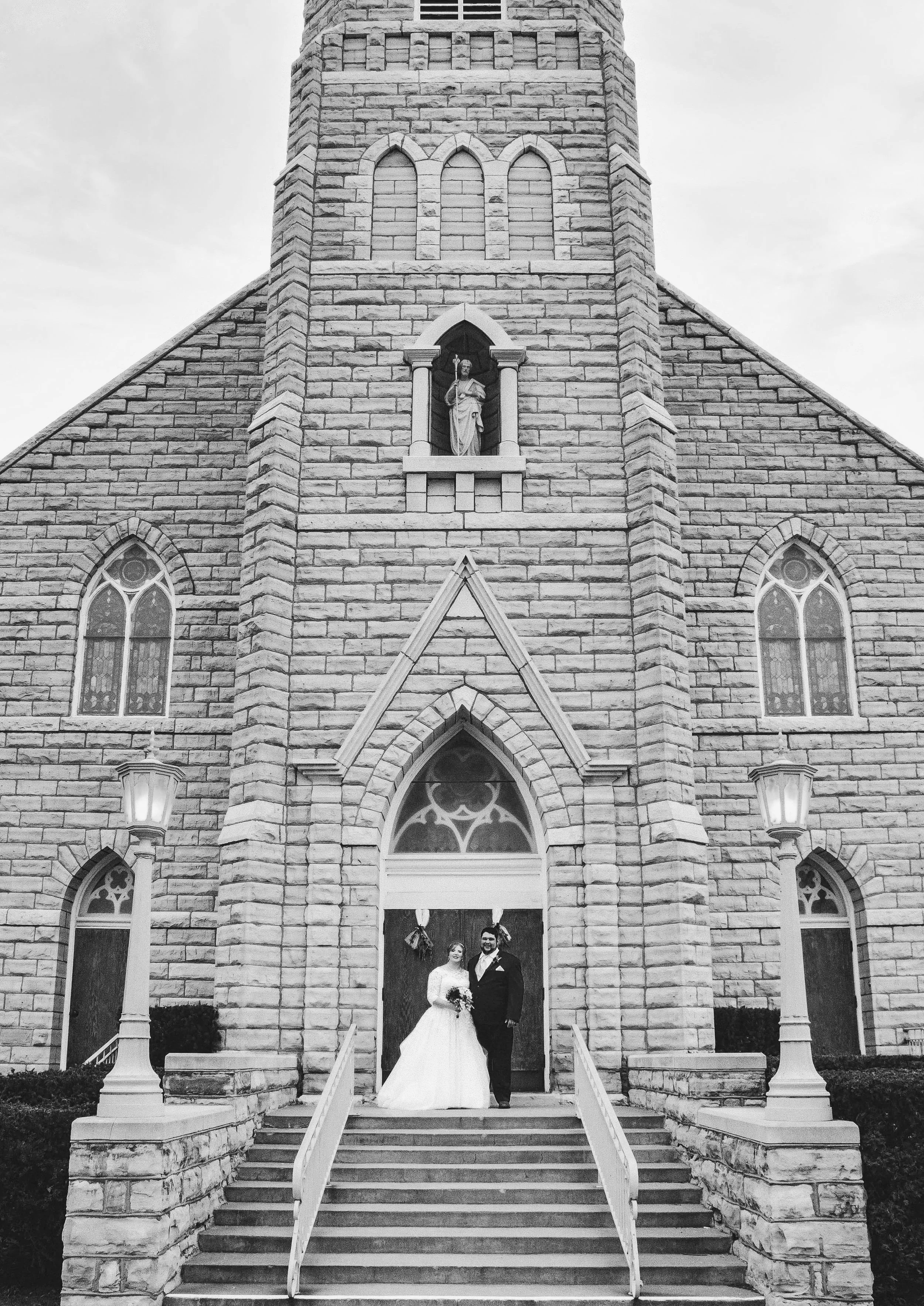 A bride and groom standing on the steps of a stone church, dressed in wedding attire, posing for a photo in front of the church entrance with gothic windows and a statue of a saint above.