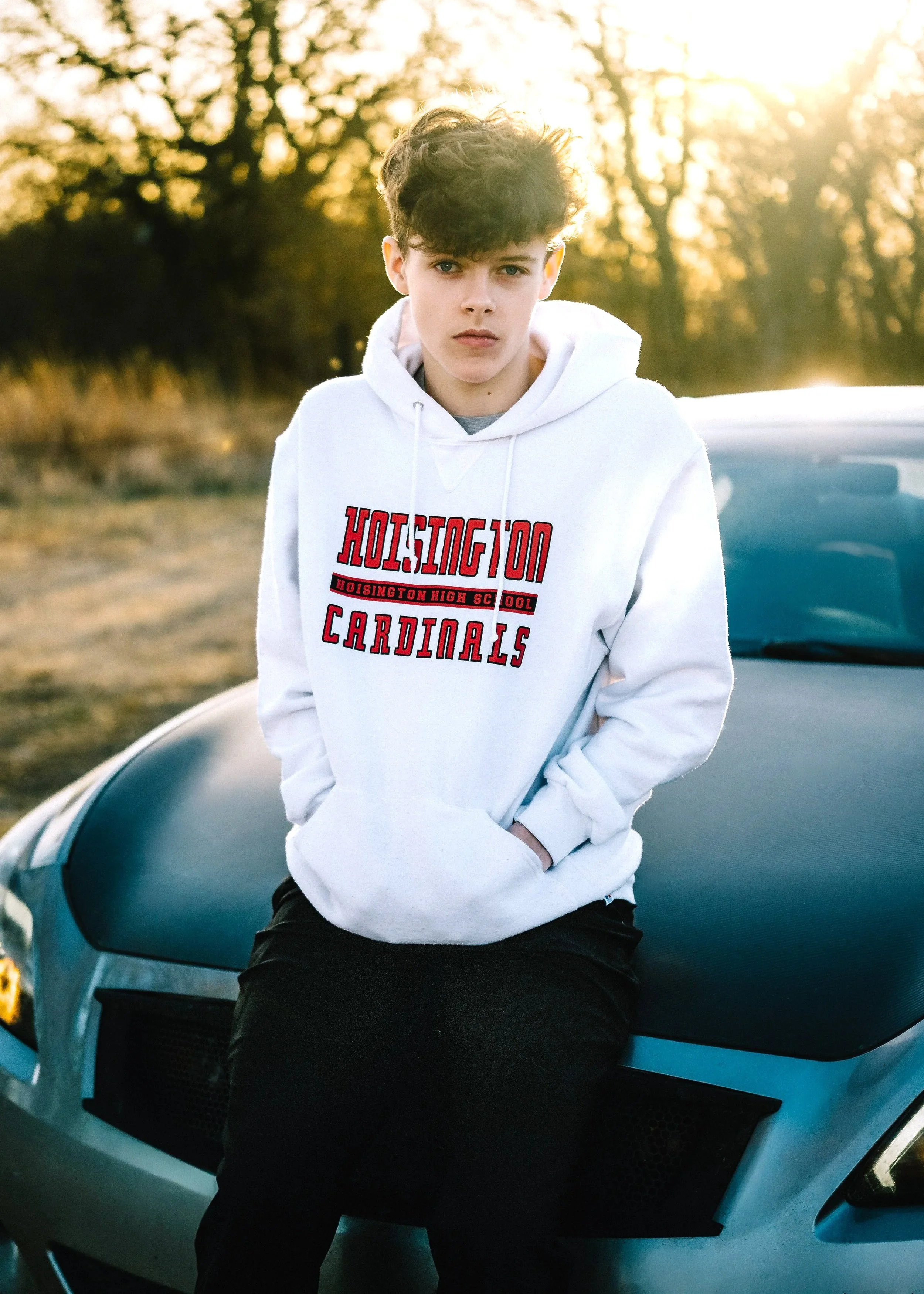 A young man with curly hair leaning against a silver sports car outdoors during sunset, wearing a white hoodie with 'Hodington Cardinals' printed on it.