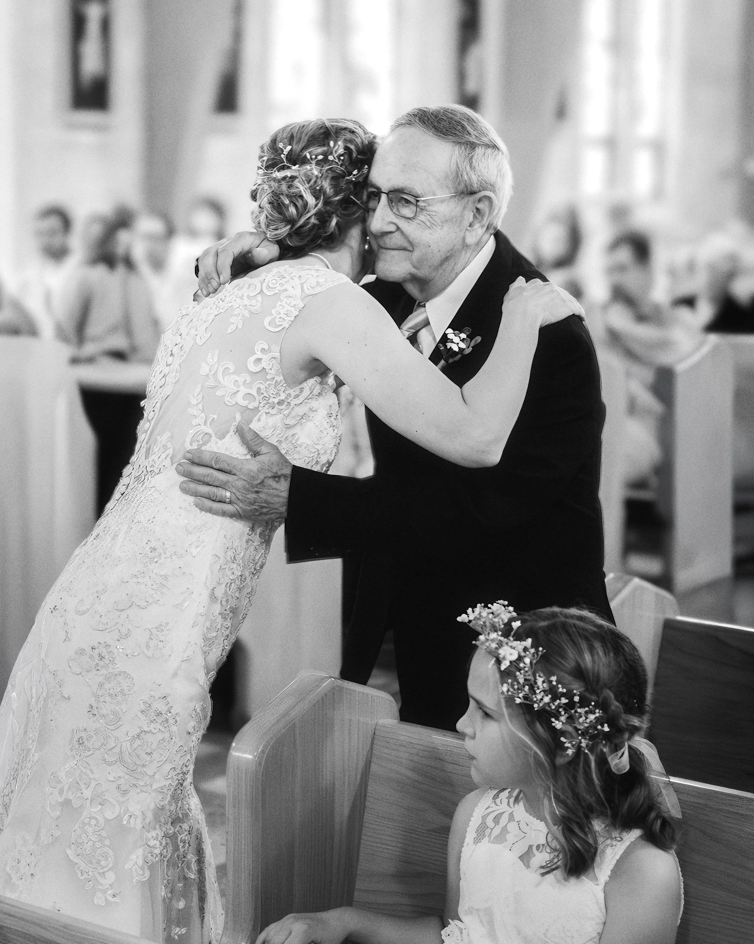 A black and white photo of a woman in a lace wedding dress hugging an older man in a suit, likely at a wedding ceremony, with a young girl with a floral headband sitting nearby.