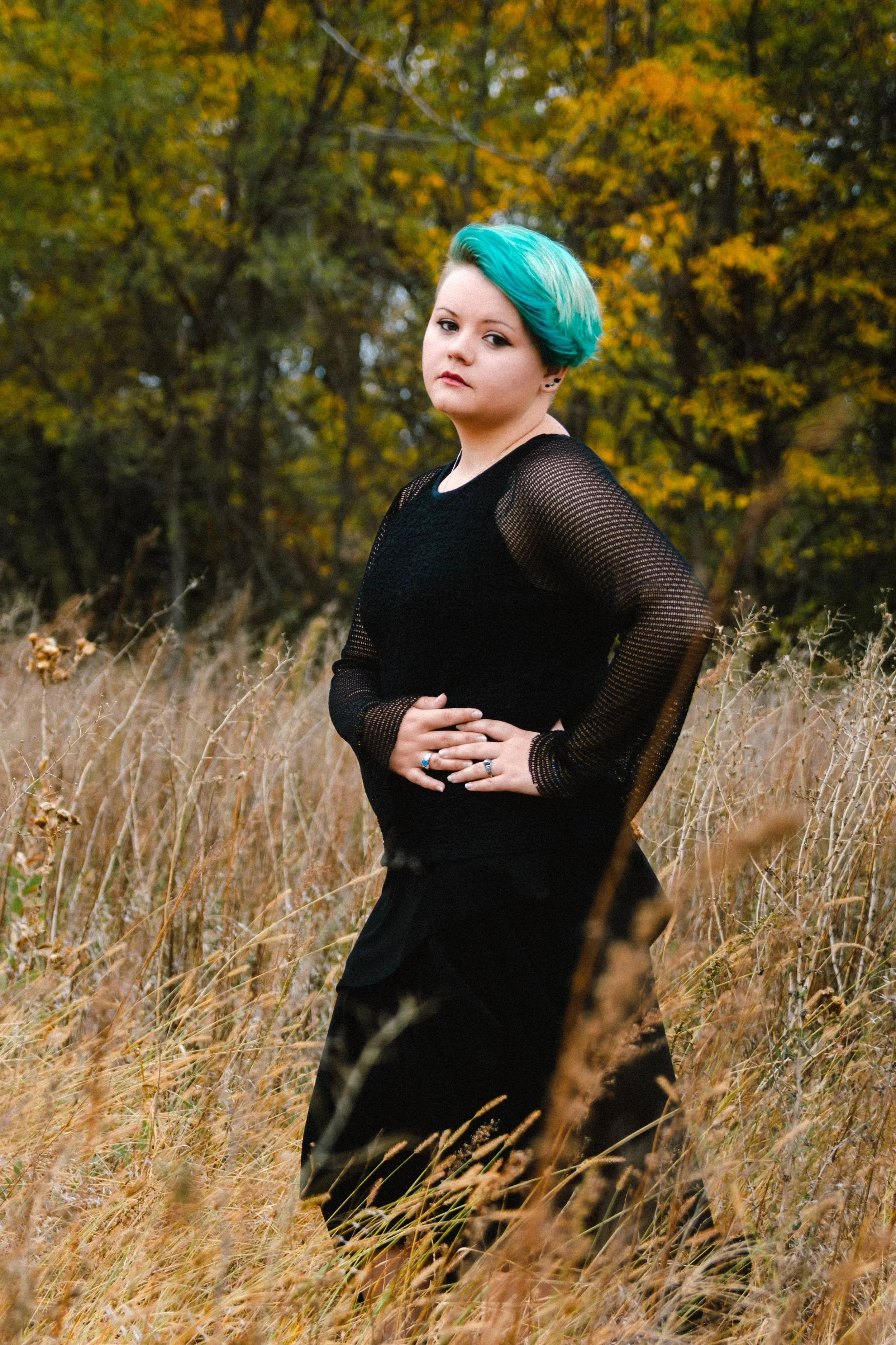 A young woman with blue and turquoise hair stands in a field of tall, dry grass during autumn, with trees showing yellow and orange leaves in the background. She is wearing a black mesh long-sleeve top and has her hands resting on her stomach.