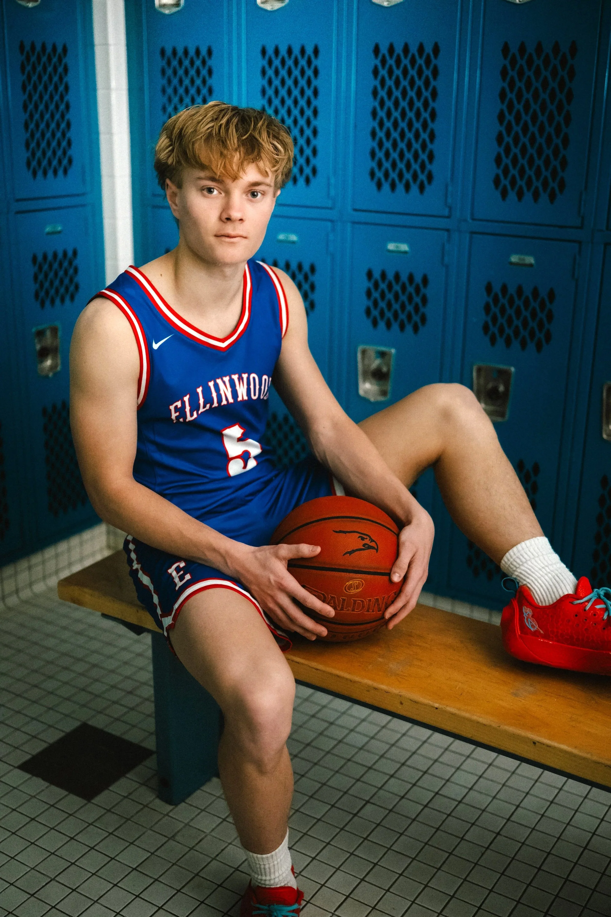 A young male basketball player sitting on a bench in a locker room, holding a basketball, wearing a blue jersey with the number 5, red and white shoes, and white socks. Blue lockers are in the background.