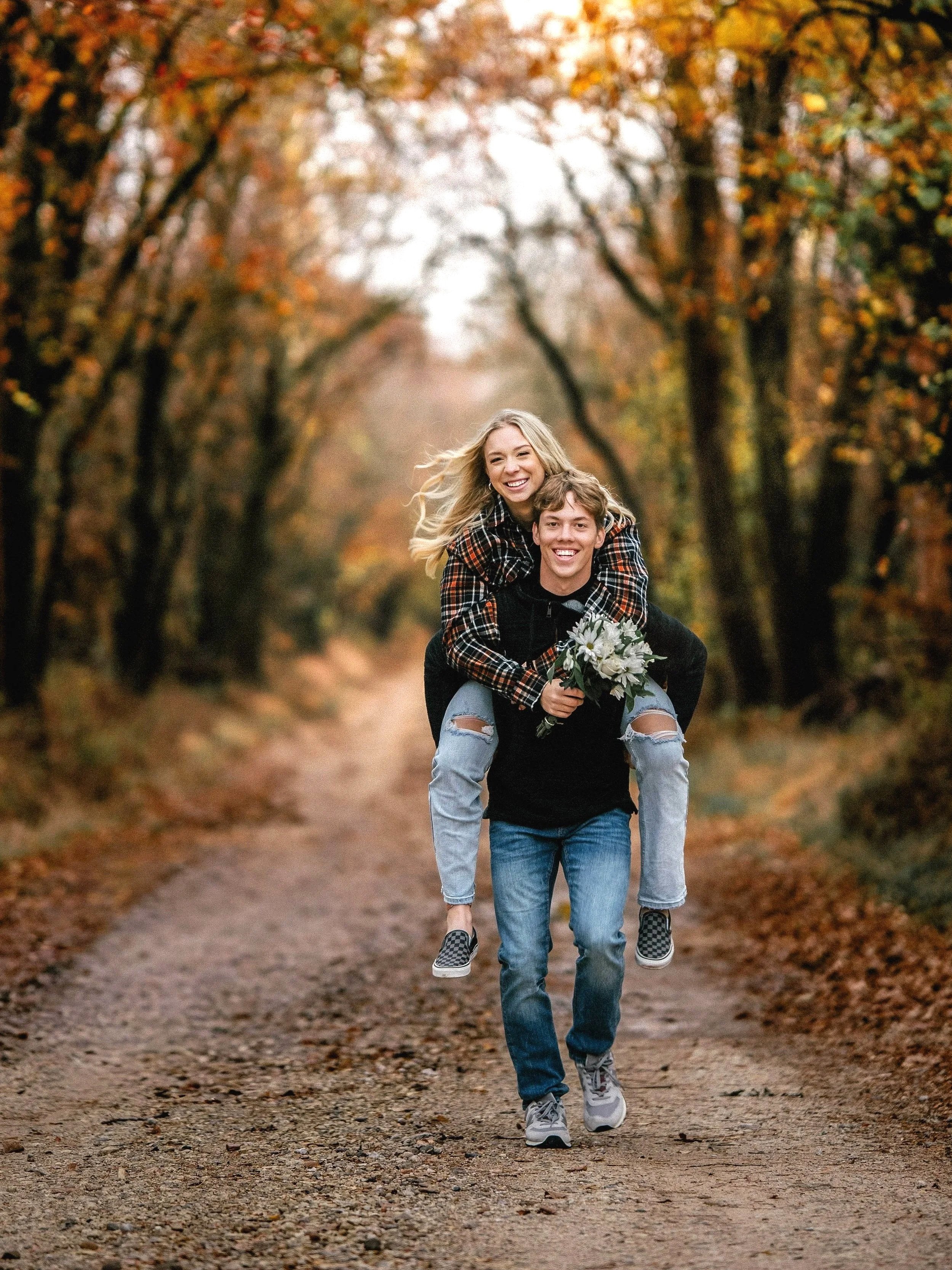 A young couple enjoying an autumn walk on a dirt path surrounded by colorful fall trees. The woman is riding on the man's back, holding a bouquet of white flowers, and both are smiling.