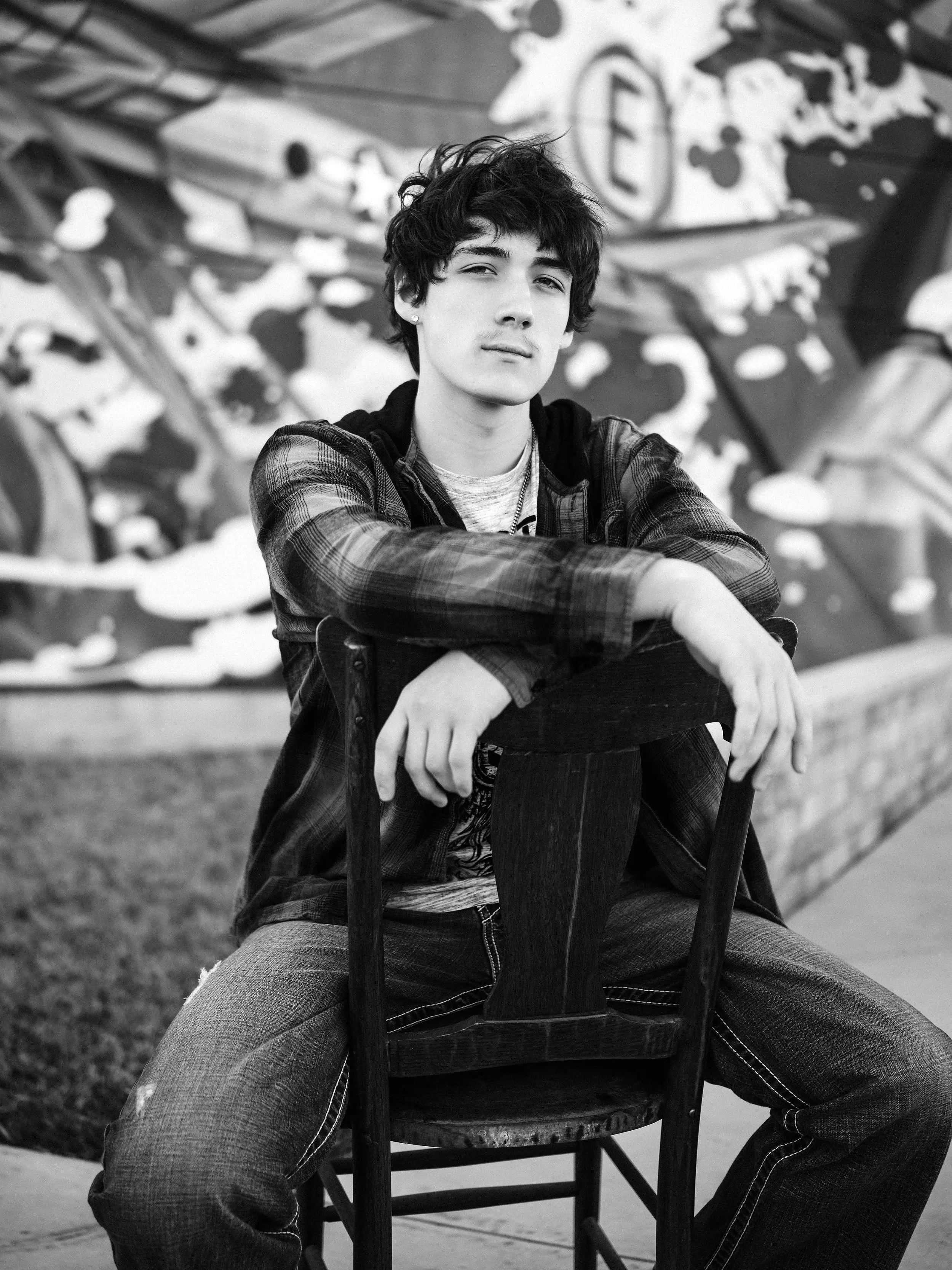 A young man with dark, tousled hair is sitting on a wooden chair outdoors, with his arm resting on the backrest. He is wearing a plaid shirt layered over a T-shirt and appears to be in front of a colorful graffiti wall.