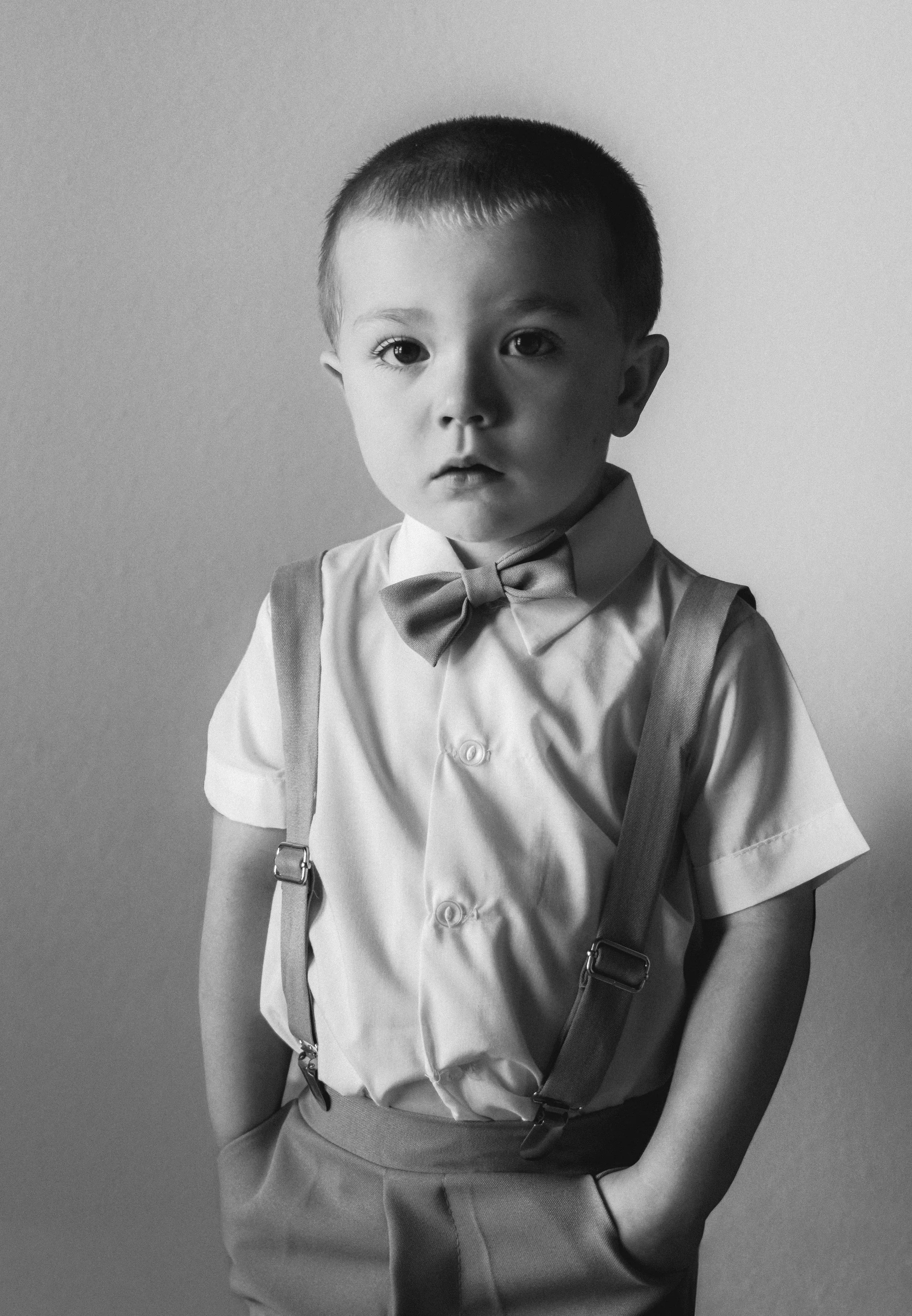 Black and white photo of a young boy dressed in a collared shirt with a bow tie, suspenders, and pants, standing against a plain background with a serious expression.