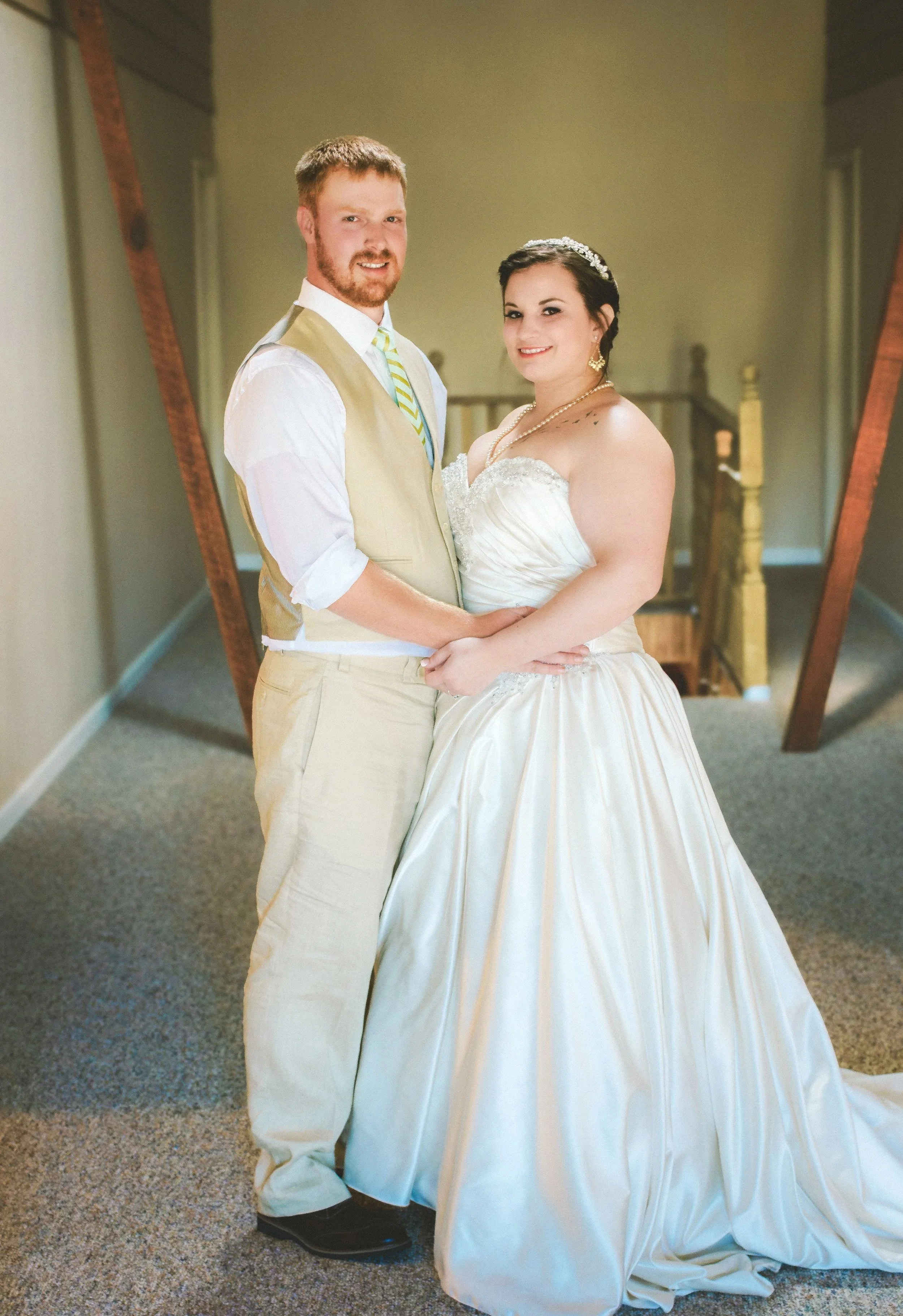 A newlywed couple standing on a staircase landing, posing for a wedding photo indoors. The bride is wearing a strapless satin wedding gown with a full skirt, and the groom is dressed in a light-colored vest and pants with a striped tie.