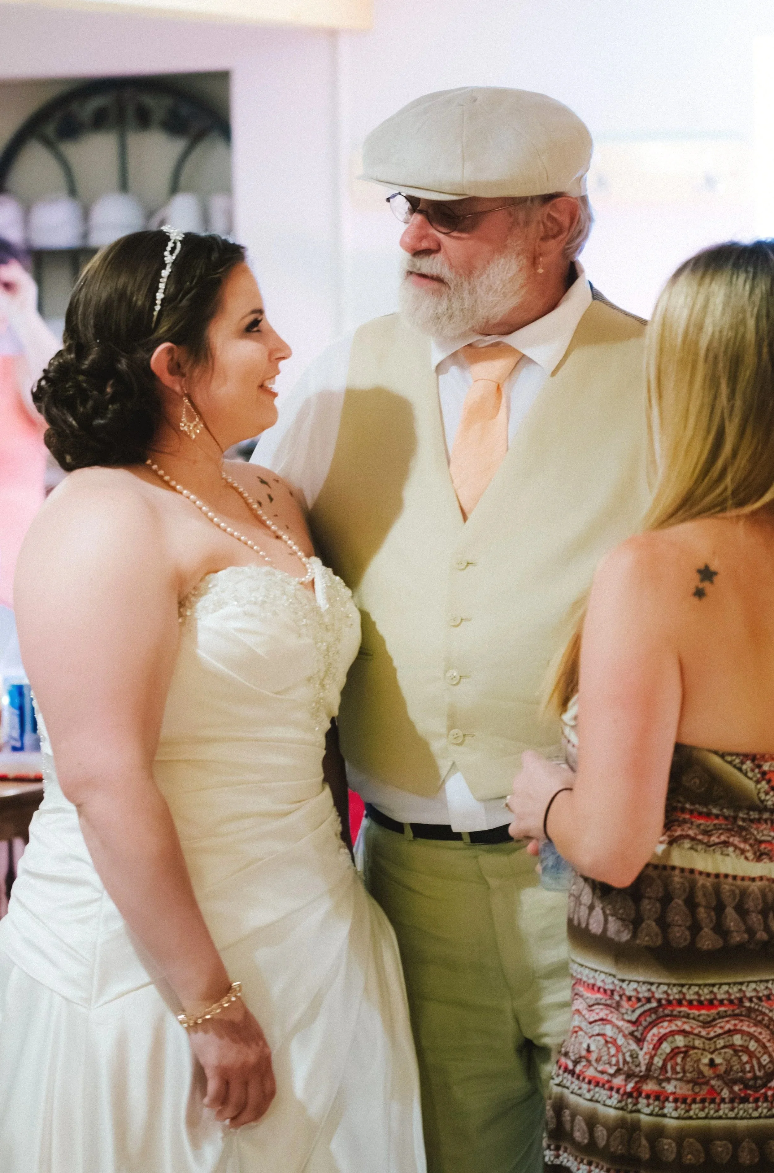A bride and an older man with glasses and a beard share a moment with two women during a celebration, likely a wedding reception, in an indoor setting.