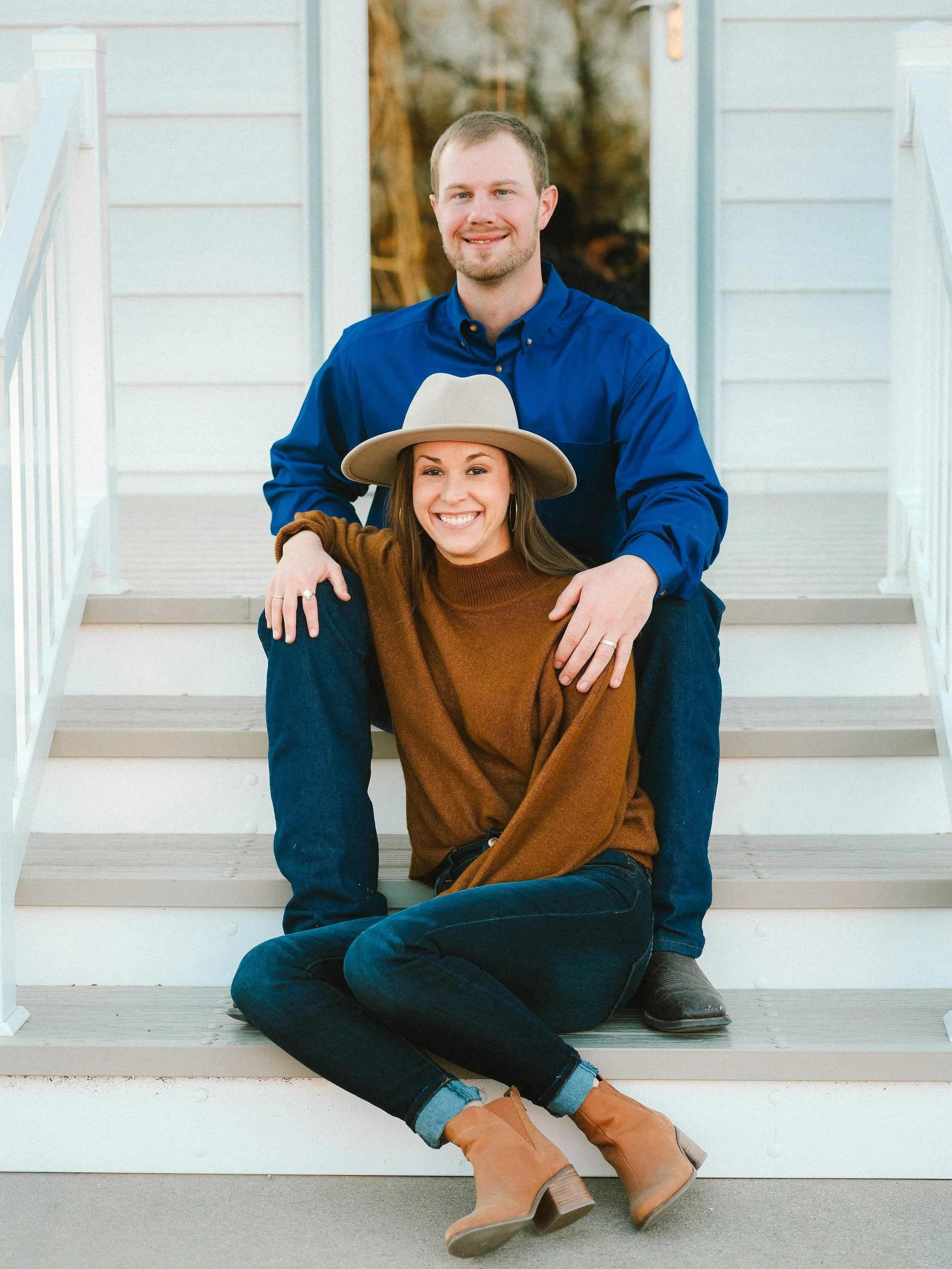 A smiling man and woman sitting on porch steps, with the woman sitting on the steps and the man kneeling behind her, both looking at the camera. The woman is wearing a tan hat, brown sweater, blue jeans, and tan ankle boots. The man is wearing a blue