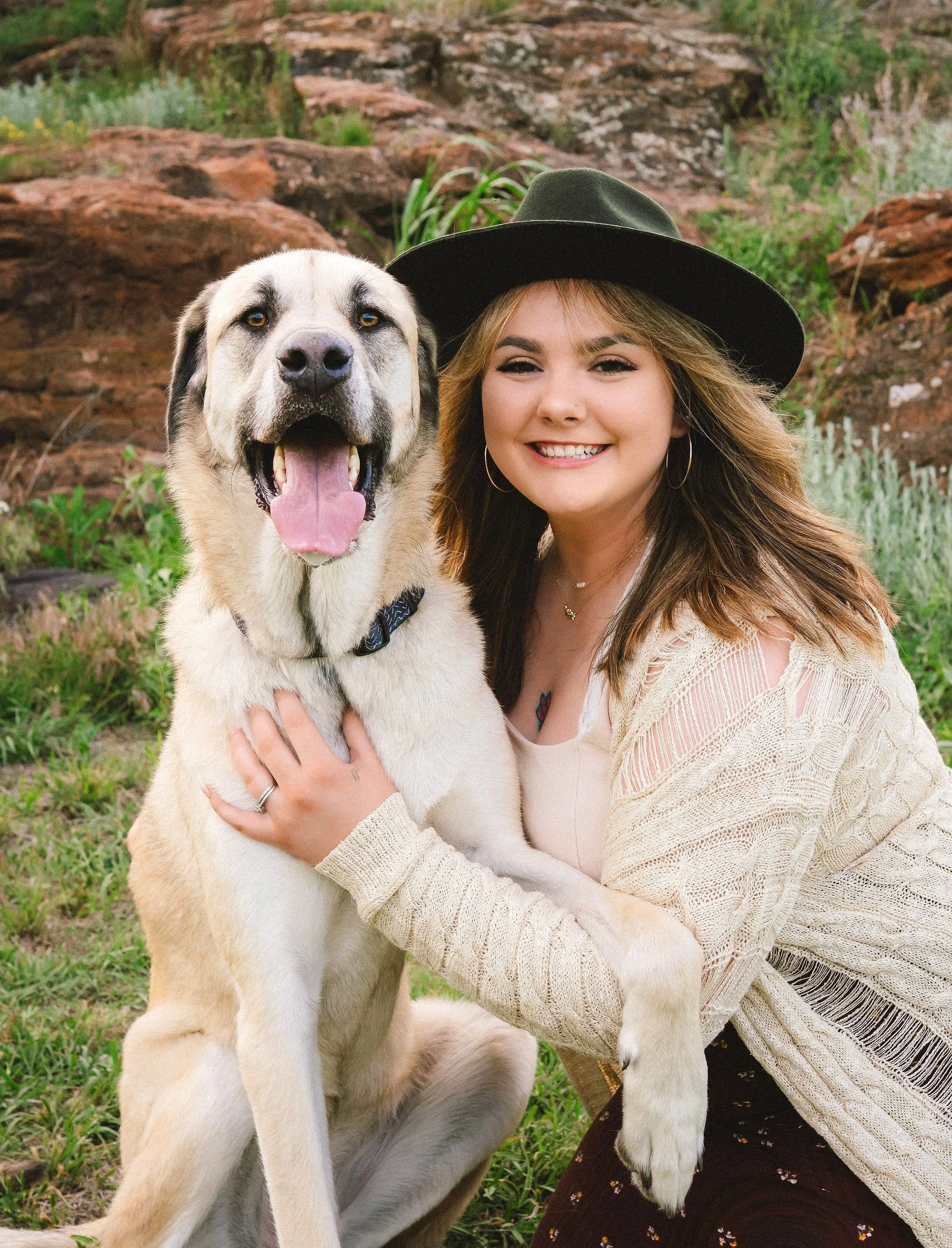 A young woman with a hat and a beige sweater smiling and hugging a large, happy dog outdoors with rocks and greenery in the background.
