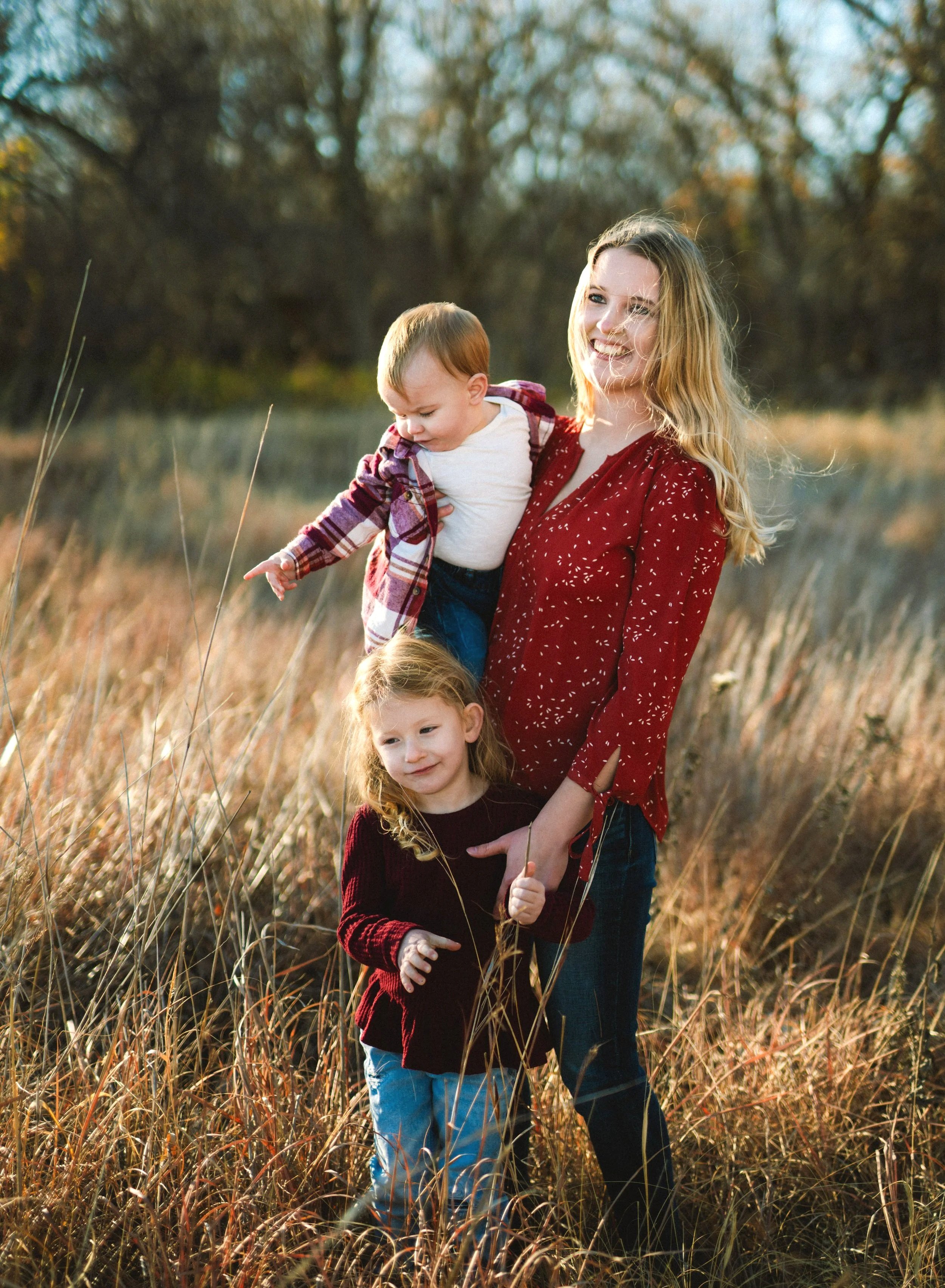 A woman with two children standing in a field of tall grass with trees in the background, enjoying a sunny day outdoors.