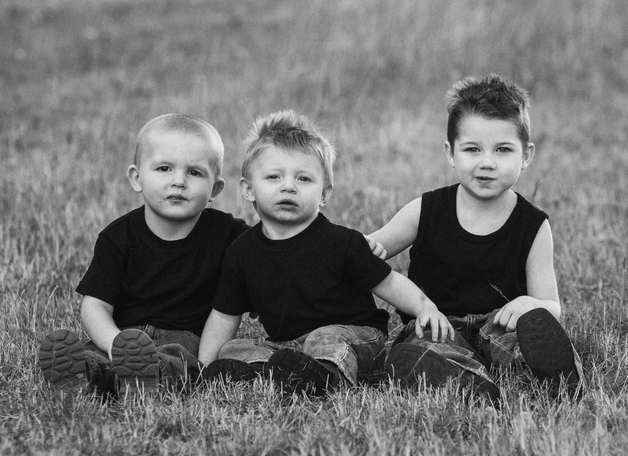 Three young boys sitting on grass, wearing black shirts and jeans, outdoors.